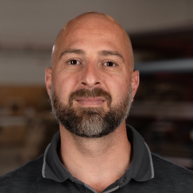 A man with a bald head, beard, and mustache, wearing a dark collared shirt, looks directly at the camera with a neutral expression. The background is blurred, suggesting an indoor setting.