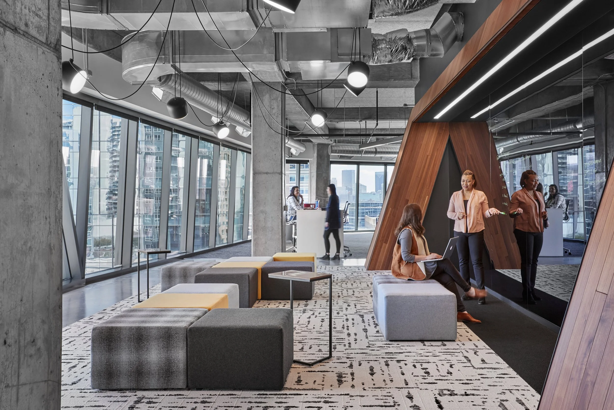 Modern office lounge area with large windows, exposed concrete walls, and industrial-style lighting. People are working and socializing, some sitting on upholstered benches, others standing and talking, with city skyscrapers visible outside.
