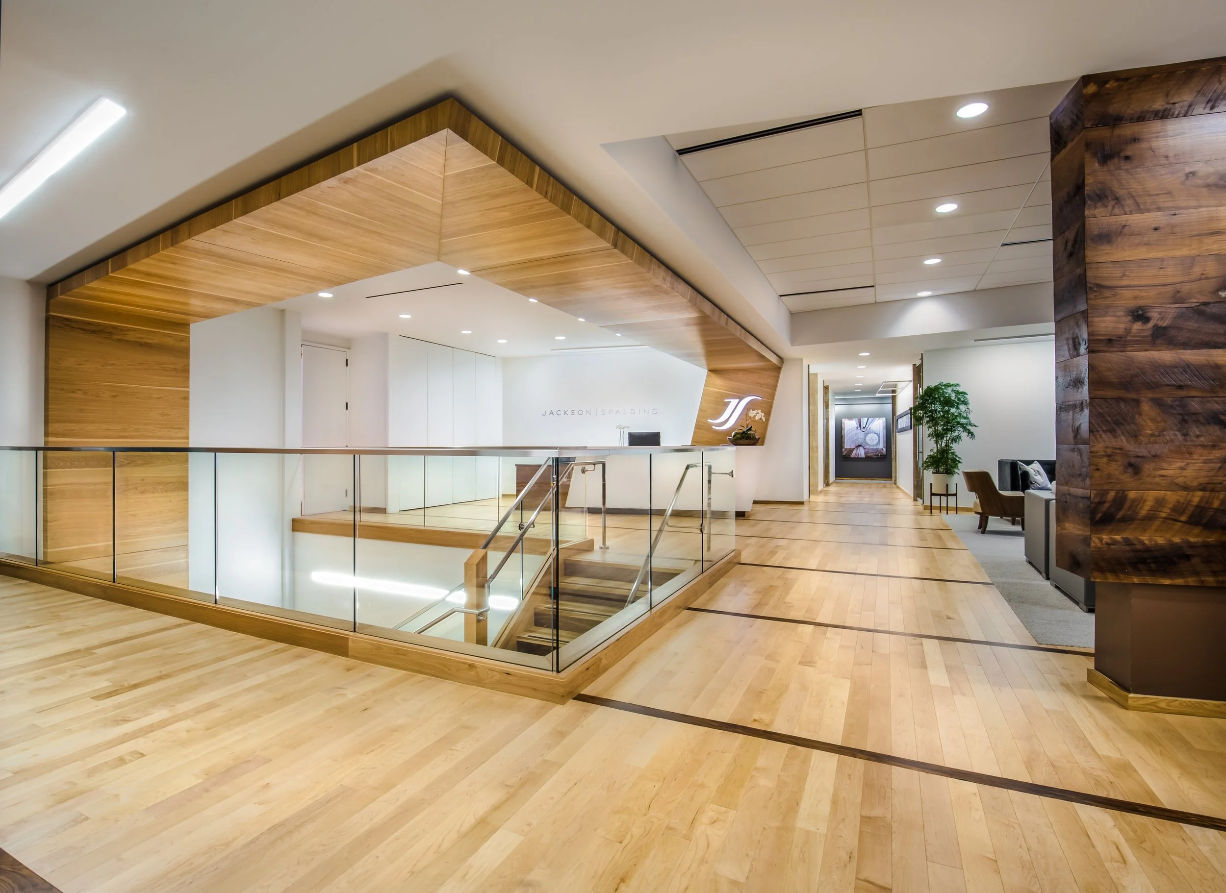 Interior of a modern office with wooden accents, glass railing, and a staircase, featuring a reception area and seating with a plant.
