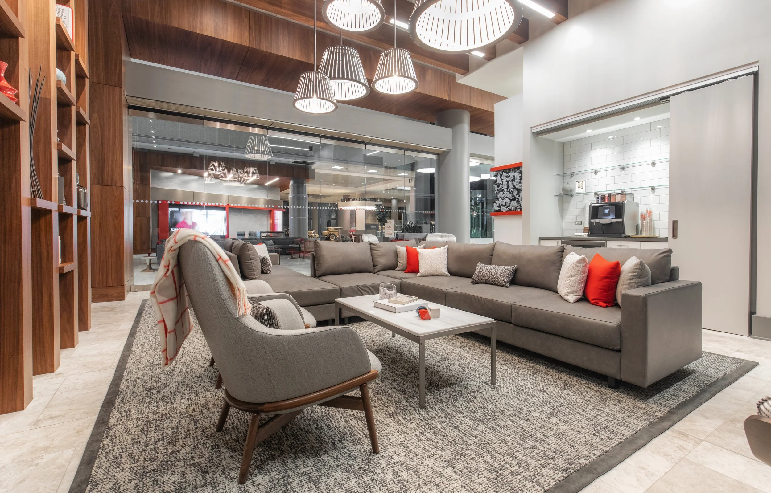 Modern hotel lobby with gray sectional sofa, patterned pillows, light gray armchair with a blanket, marble coffee table, wood-paneled walls, and hanging pendant lights.