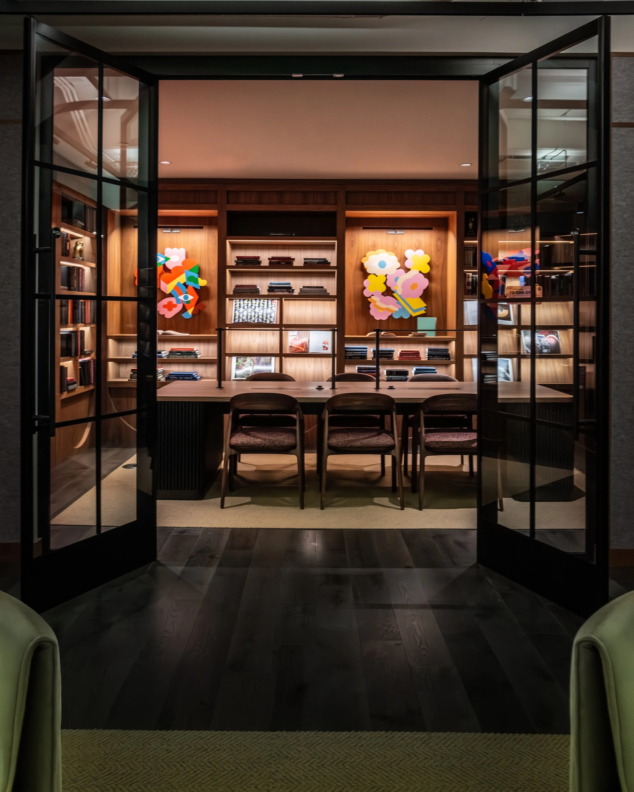 A meeting room with a wooden table and six chairs, viewed through black metal framed glass doors. The back wall has built-in wooden bookshelves with books and colorful abstract wall art.