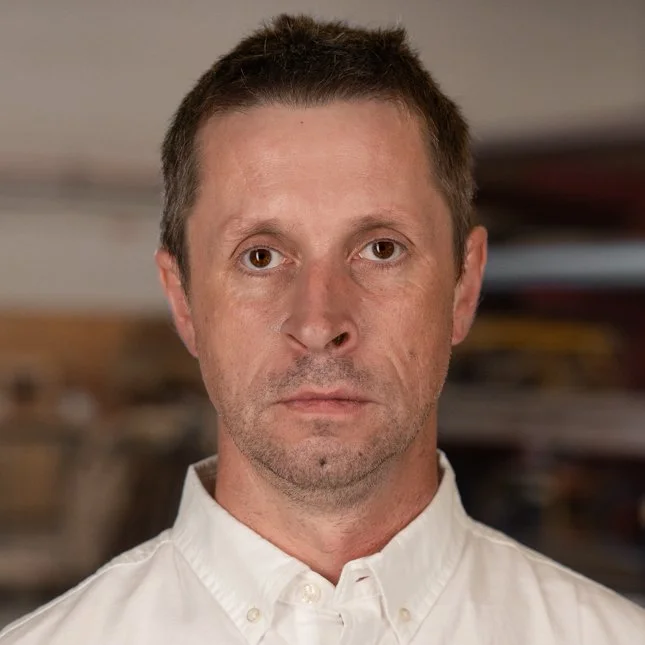 A close-up of a middle-aged man with short brown hair, wearing a white collared shirt, looking directly at the camera with a neutral expression.