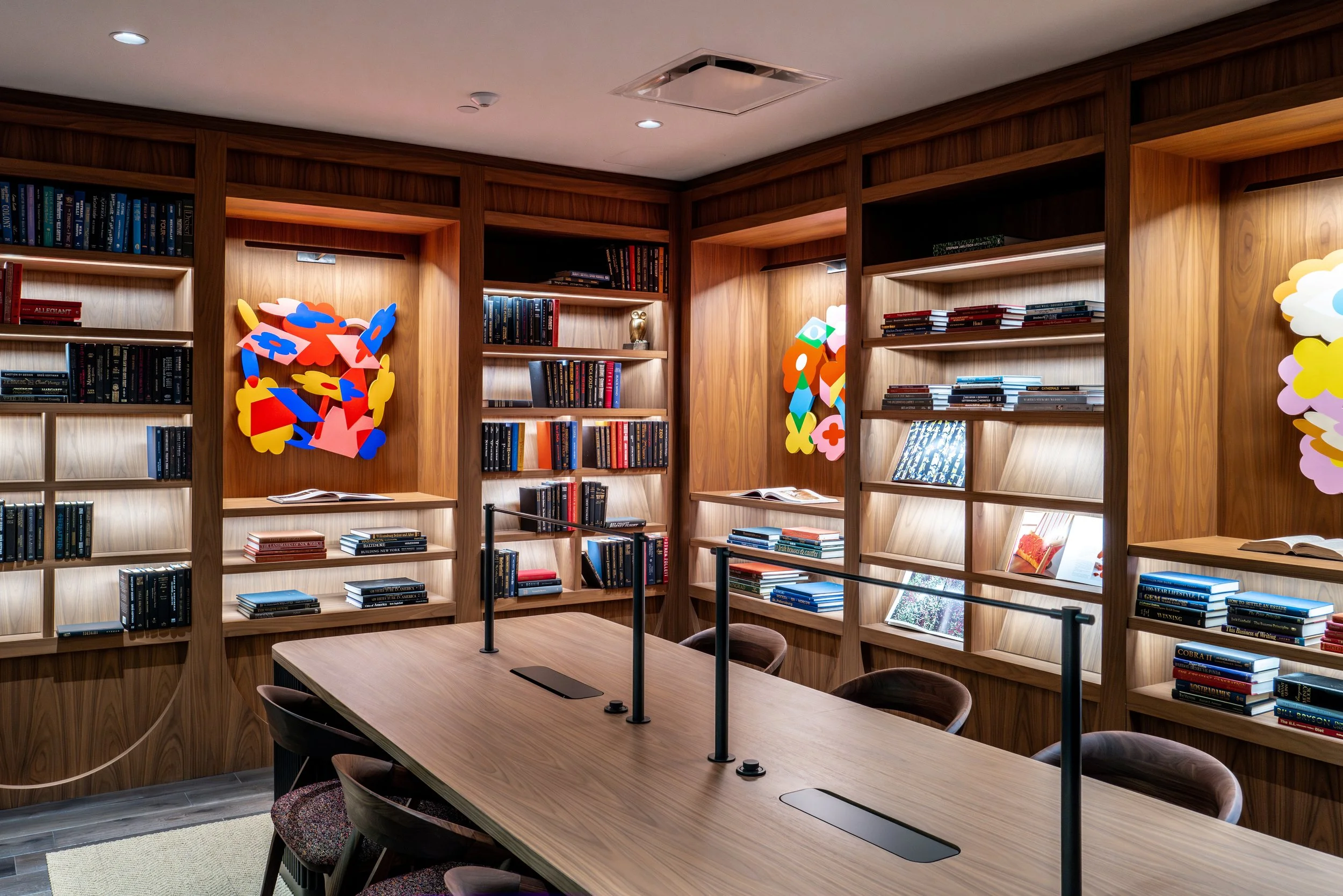 A library room with wooden bookshelves filled with books, colorful abstract art decorations on the shelves, and a large wooden table with chairs for reading or meetings.
