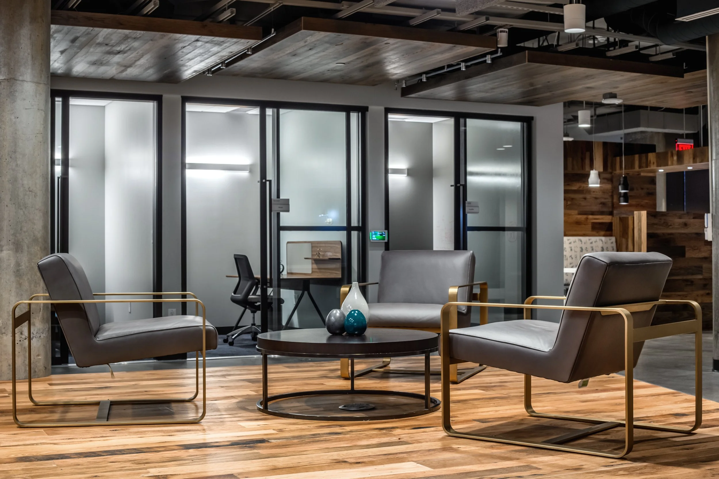 Modern office lounge area with three gray chairs with gold frames around a round black coffee table, placed on wood flooring, with frosted glass partitions and a wooden accent wall in the background.