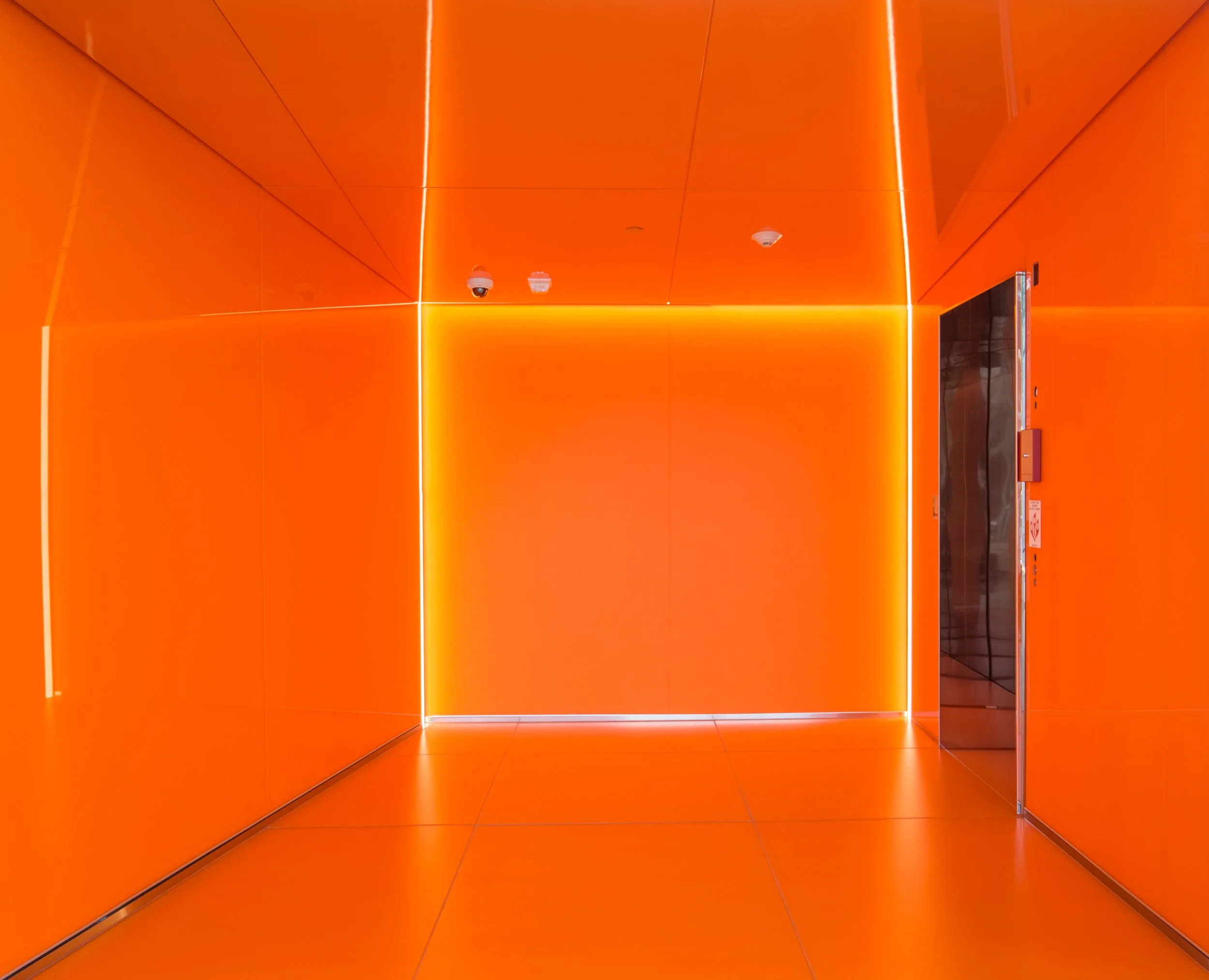 A bright orange elevator interior with reflective metal elevator door on the right side, small security cameras on the ceiling, and orange illuminated walls and ceiling with geometric panels.
