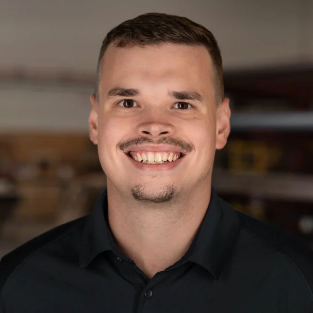 A man with short brown hair, a mustache and goatee, smiling, wearing a dark collared shirt, in an indoor setting with blurred background.