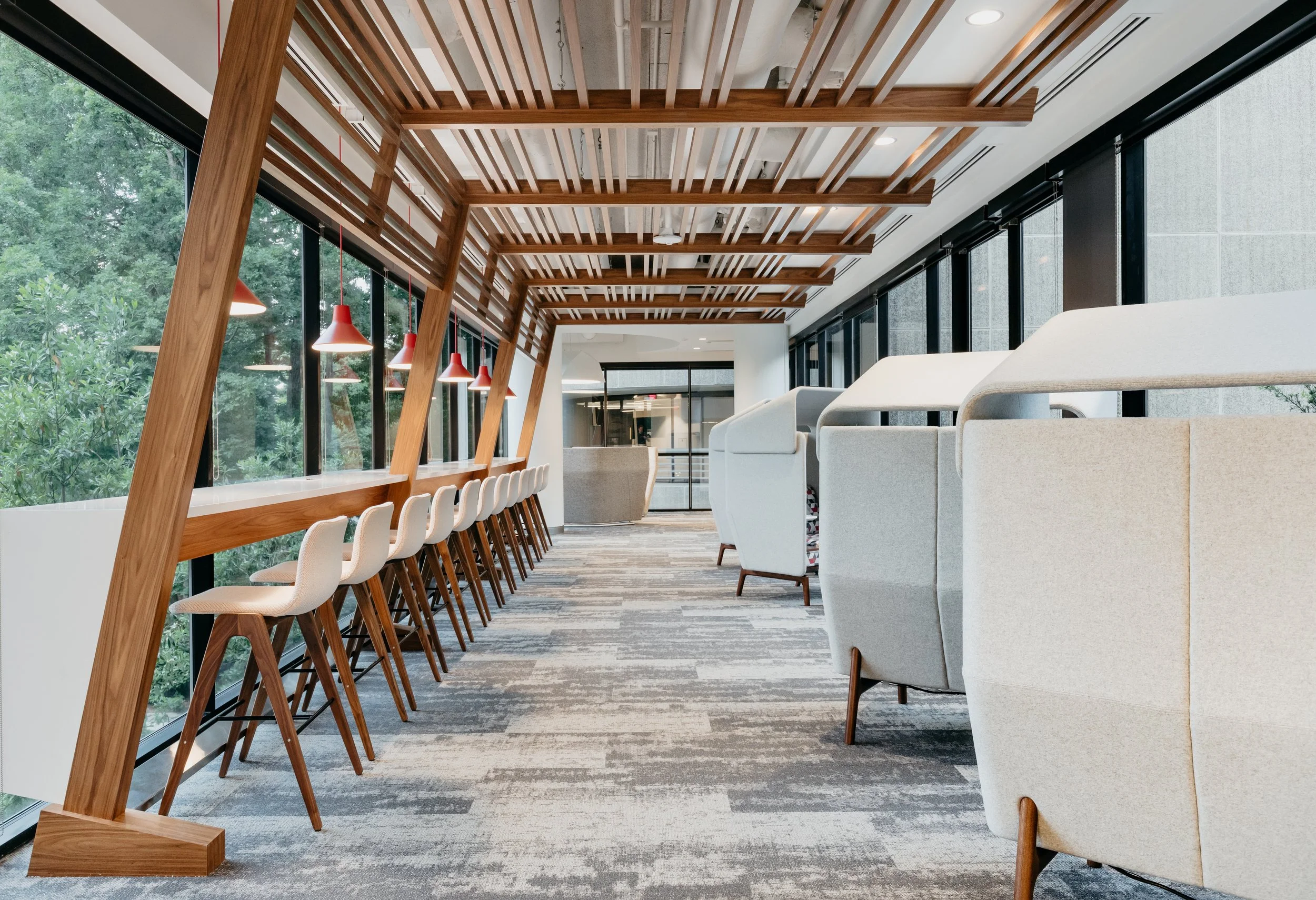 Modern interior with a row of white bar stools along a windowed wall on the left, and light-colored armchairs on the right, with a wooden slatted ceiling and large windows showing green trees outside.