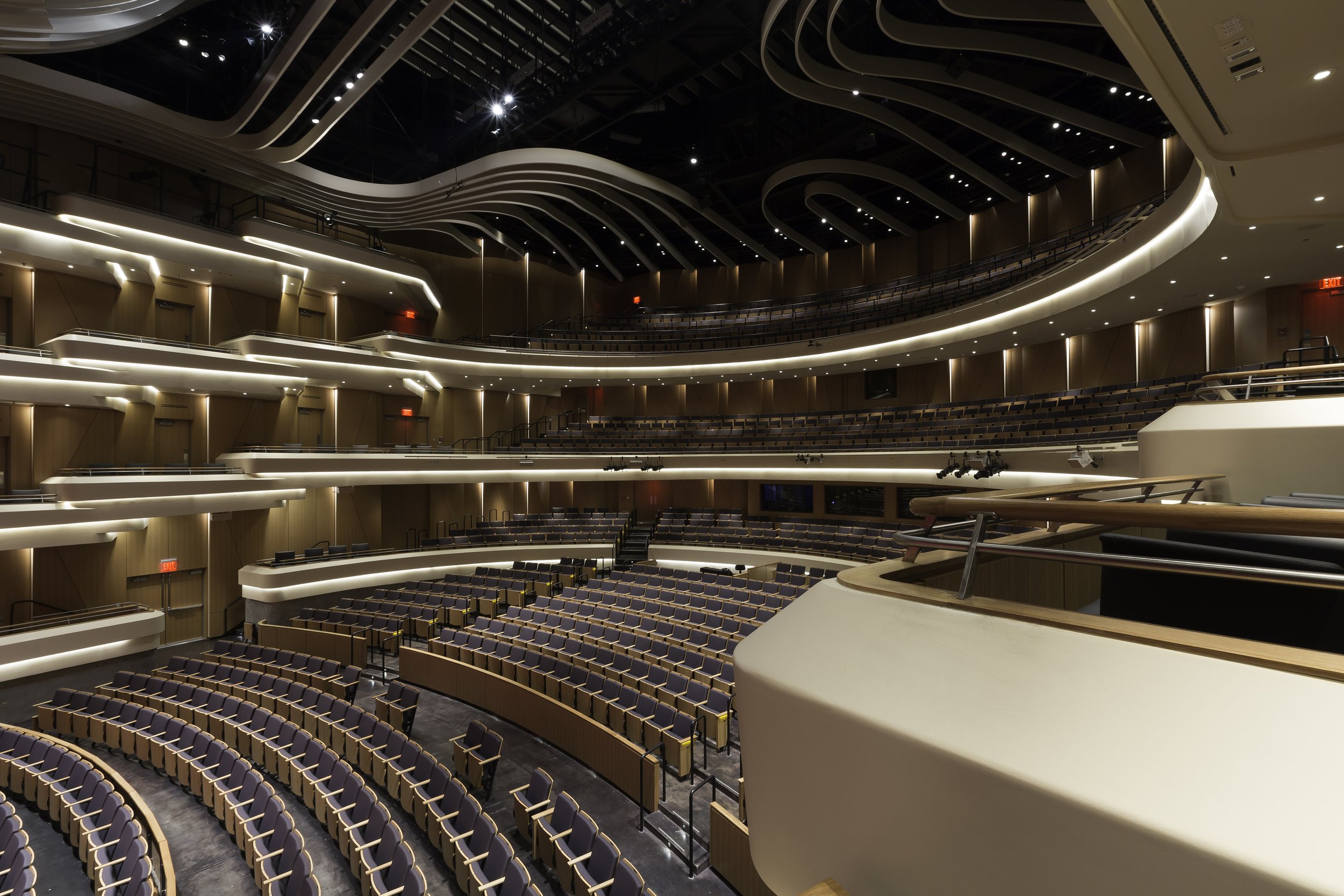 Interior view of a modern theater or auditorium with multiple tiers of seating, illuminated by soft lighting, featuring a balcony and a spacious stage area.