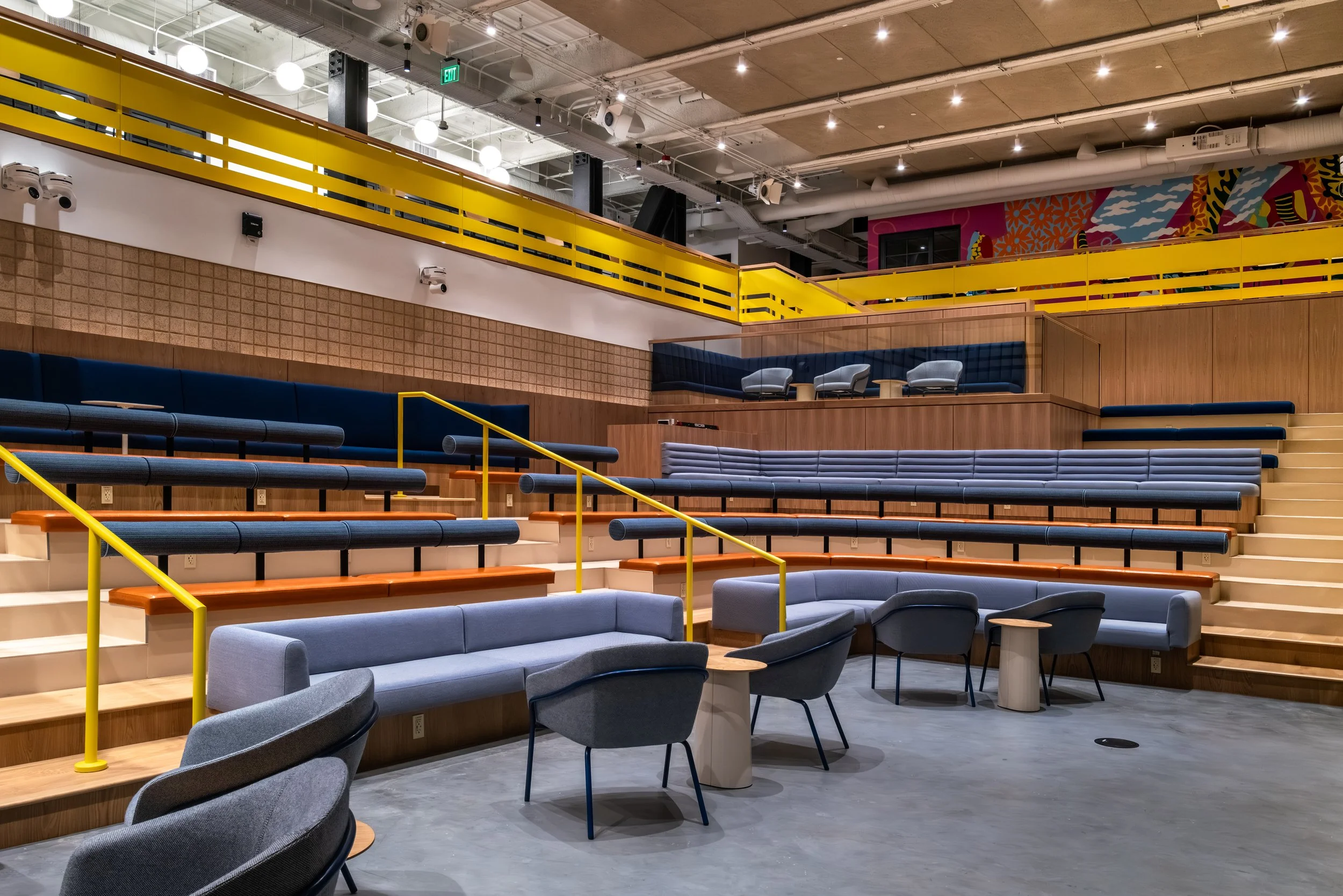 Modern auditorium or lounge with tiered seating, upholstered chairs, and wooden accents, illuminated by ceiling lights.