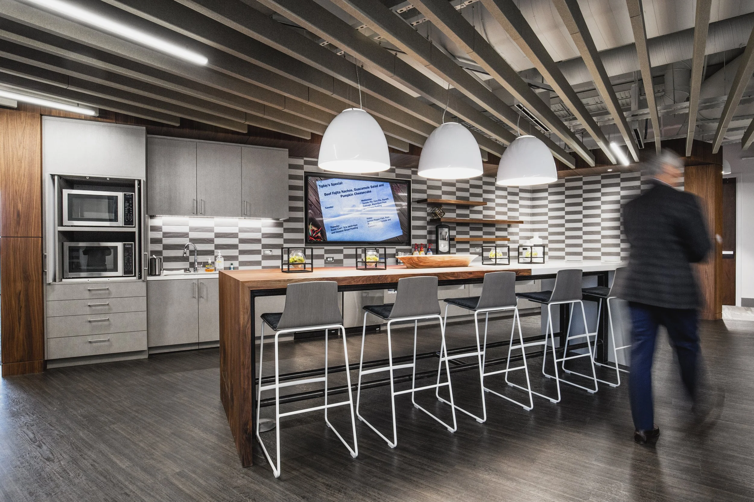 Modern office kitchen with a high island counter, bar stools, a wall-mounted display screen, and a man in a suit walking by.