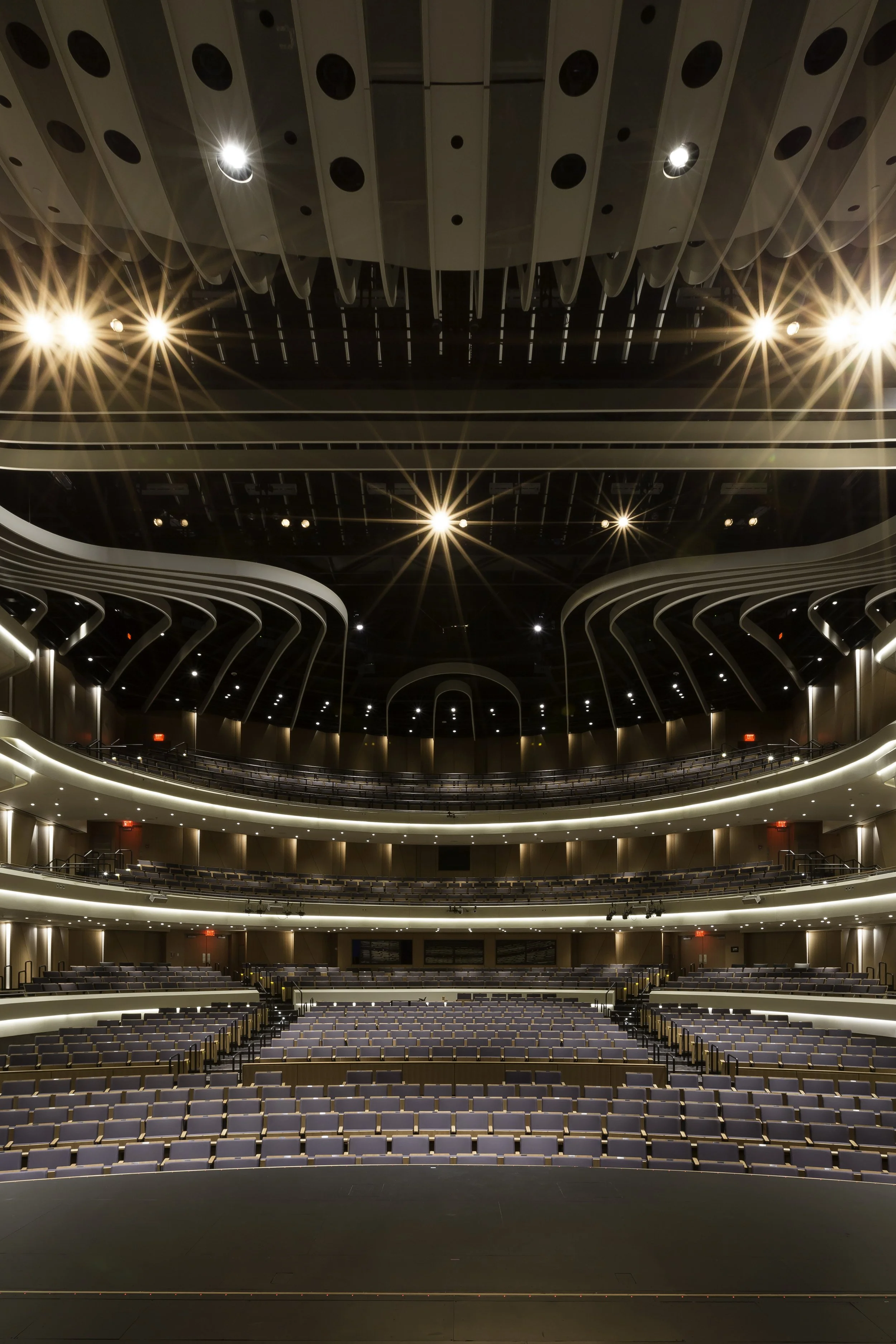 Interior of a modern theater or auditorium with rows of seats, a stage at the front, and multiple levels of balconies under a decorated ceiling, illuminated with bright stage or ceiling lights.