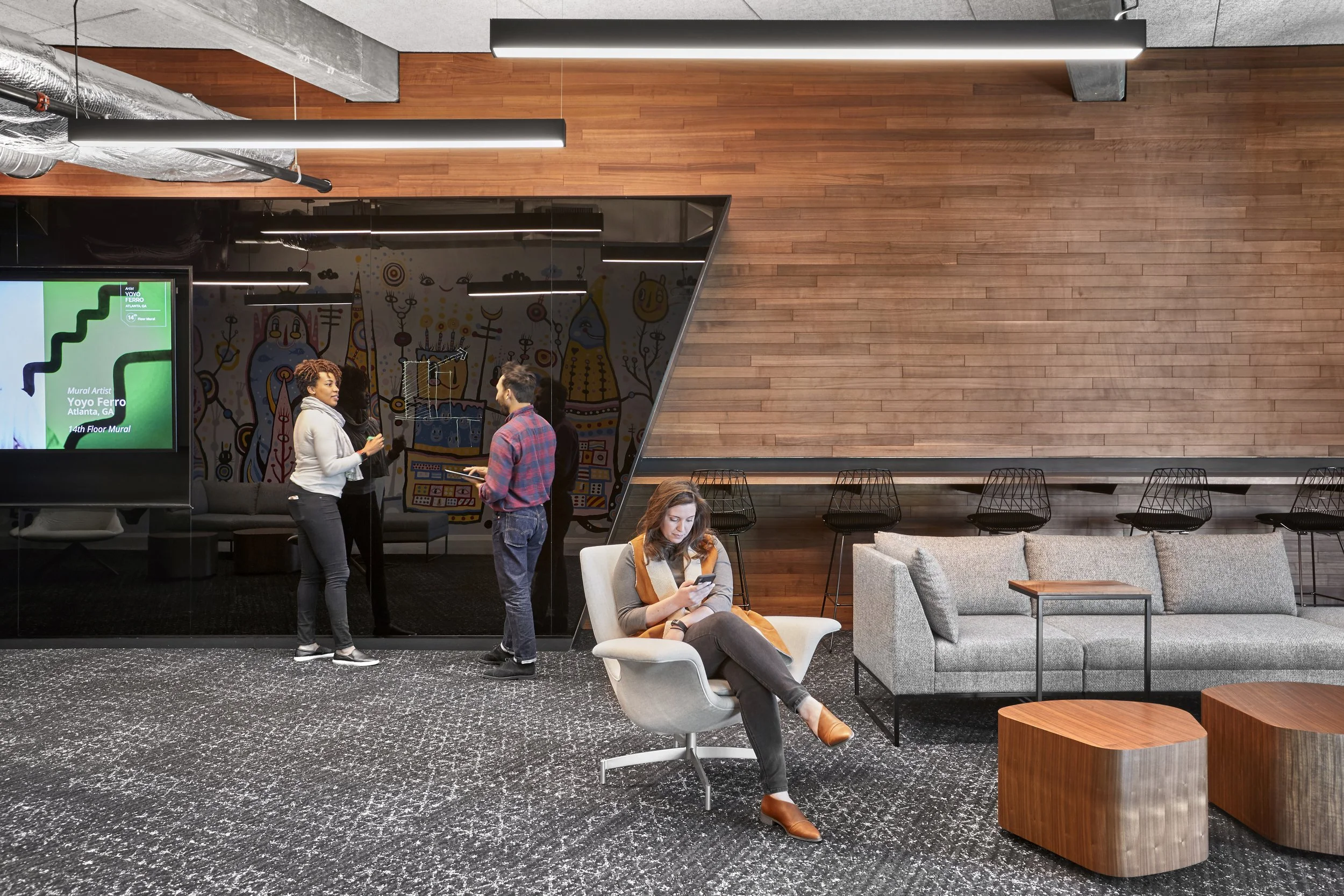 Modern office lounge area with a woman sitting on a white swivel chair looking at her phone, a gray sofa, wooden tables, and a group of people standing and talking near a glass wall with colorful mural artwork.