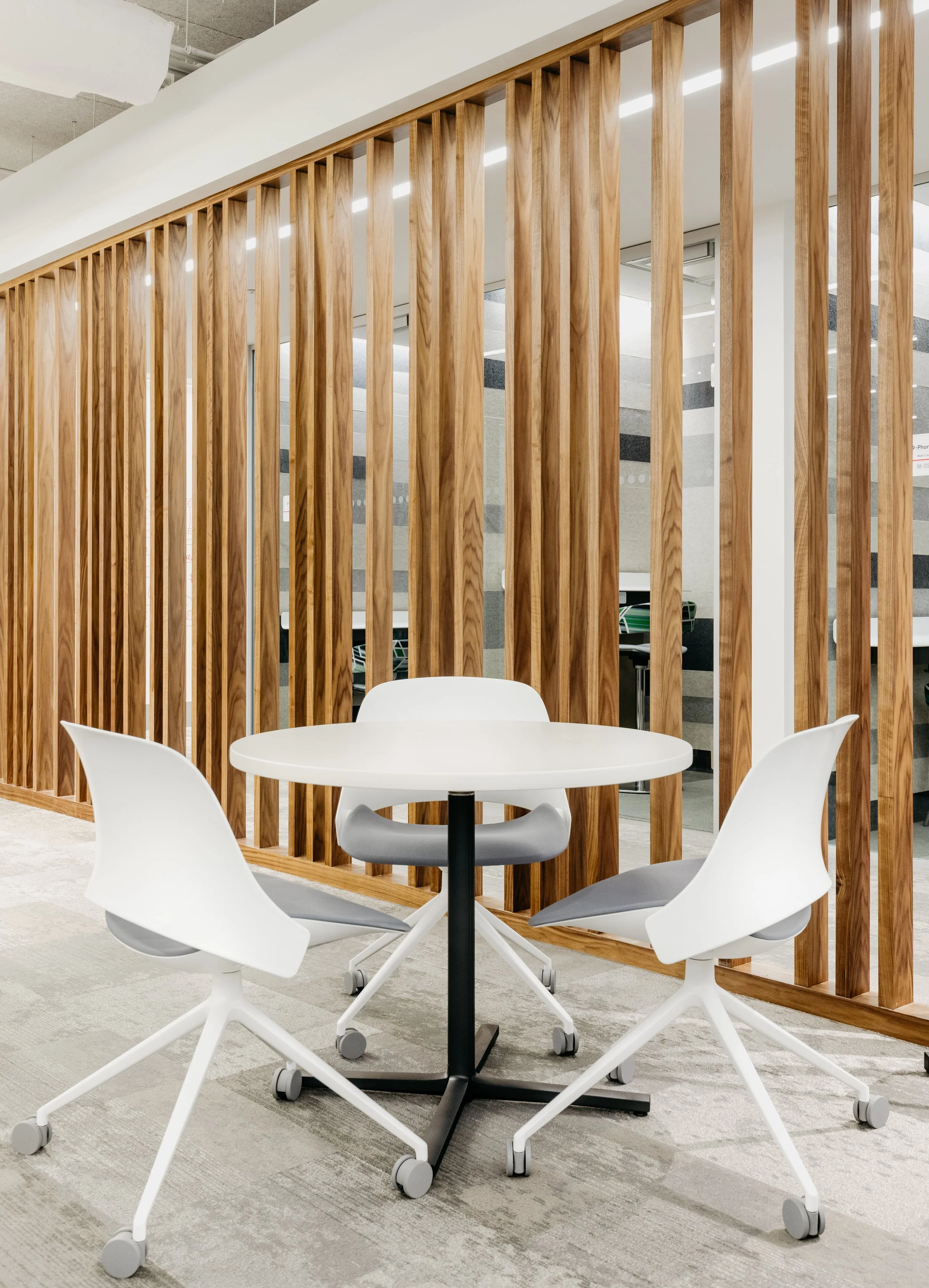 Modern office space with a white round table and three white chairs with gray cushions, separated by a wooden slat partition.