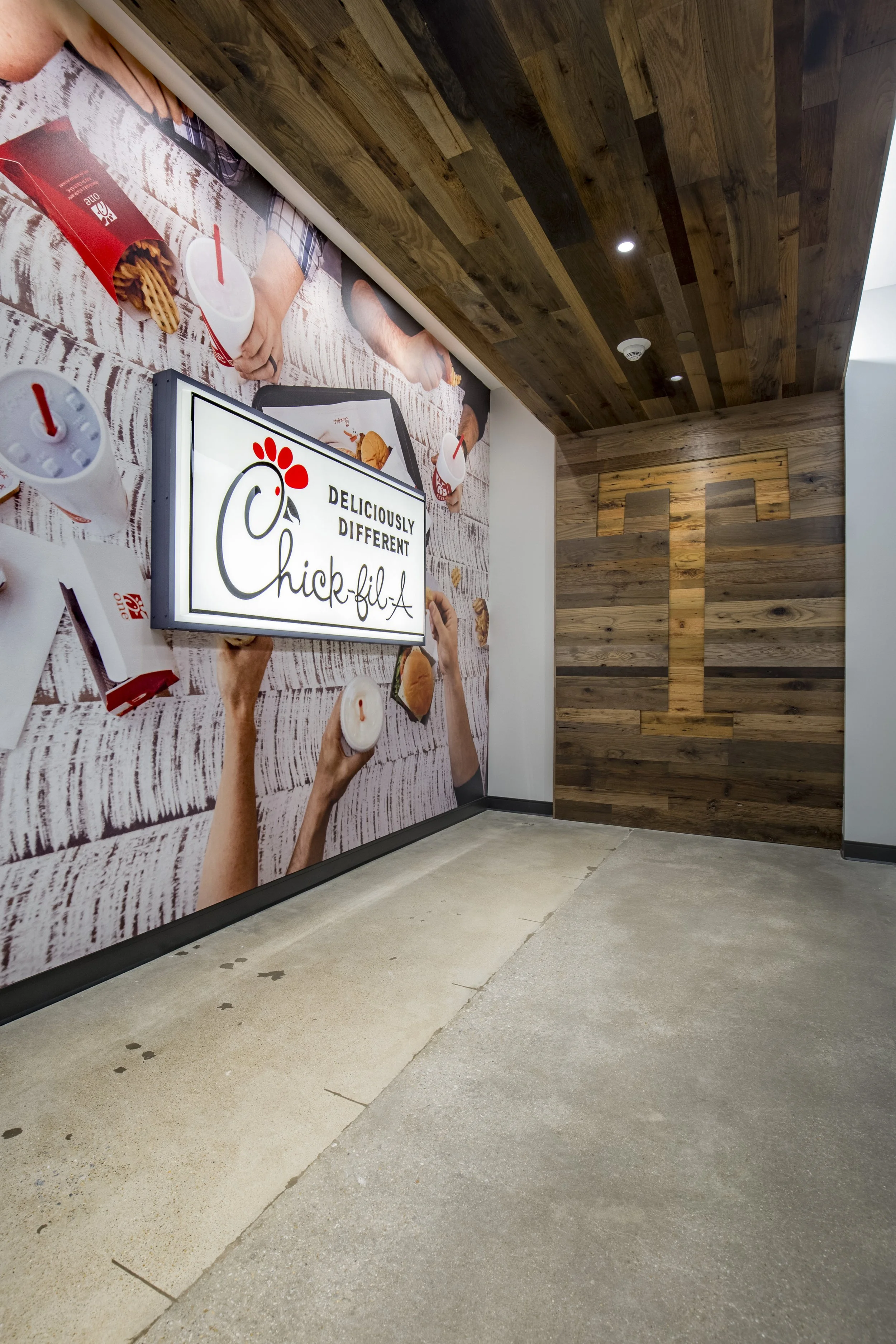 The image shows an interior wall of a Chick-fil-A restaurant with a large advertisement featuring people holding cups and food, a prominent illuminated Chick-fil-A logo with the slogan 'Deliciously Different,' and a wooden accent wall with a stylized