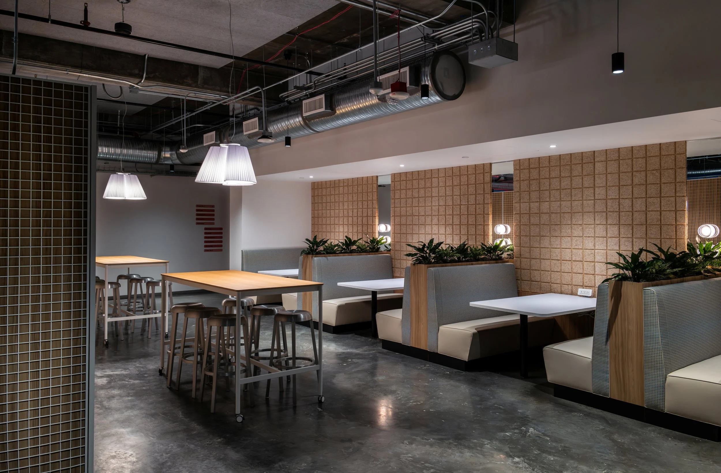 Interior of a modern, minimalist restaurant or café with booth seating, small tables, and pendant lighting, featuring exposed ceiling ducts and a concrete floor.