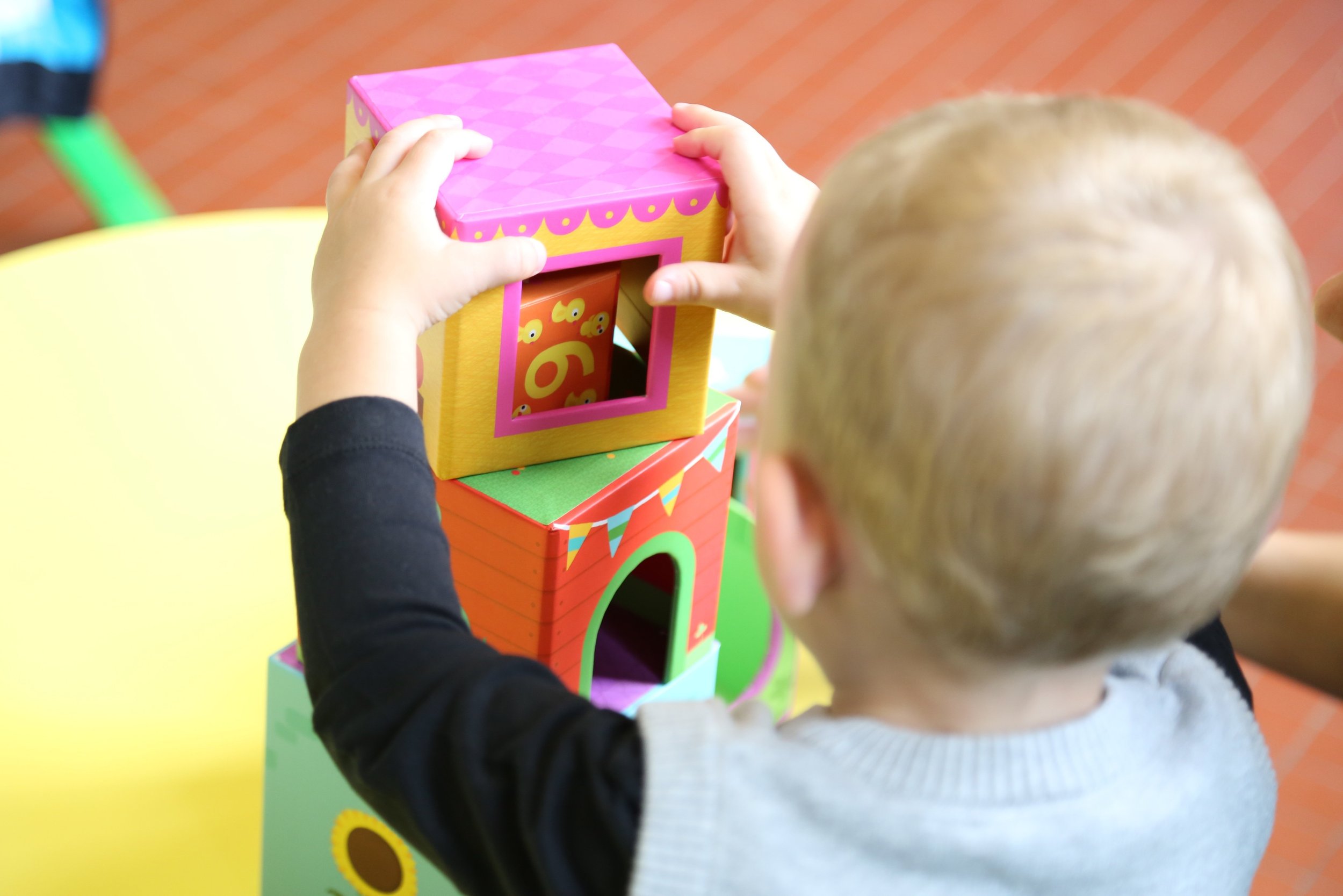 A young child with blond hair playing with colorful wooden blocks that are designed like a small house. The child is stacking a pink and yellow block on top of the structure. The background shows a yellow table and a floor with an orange pattern.