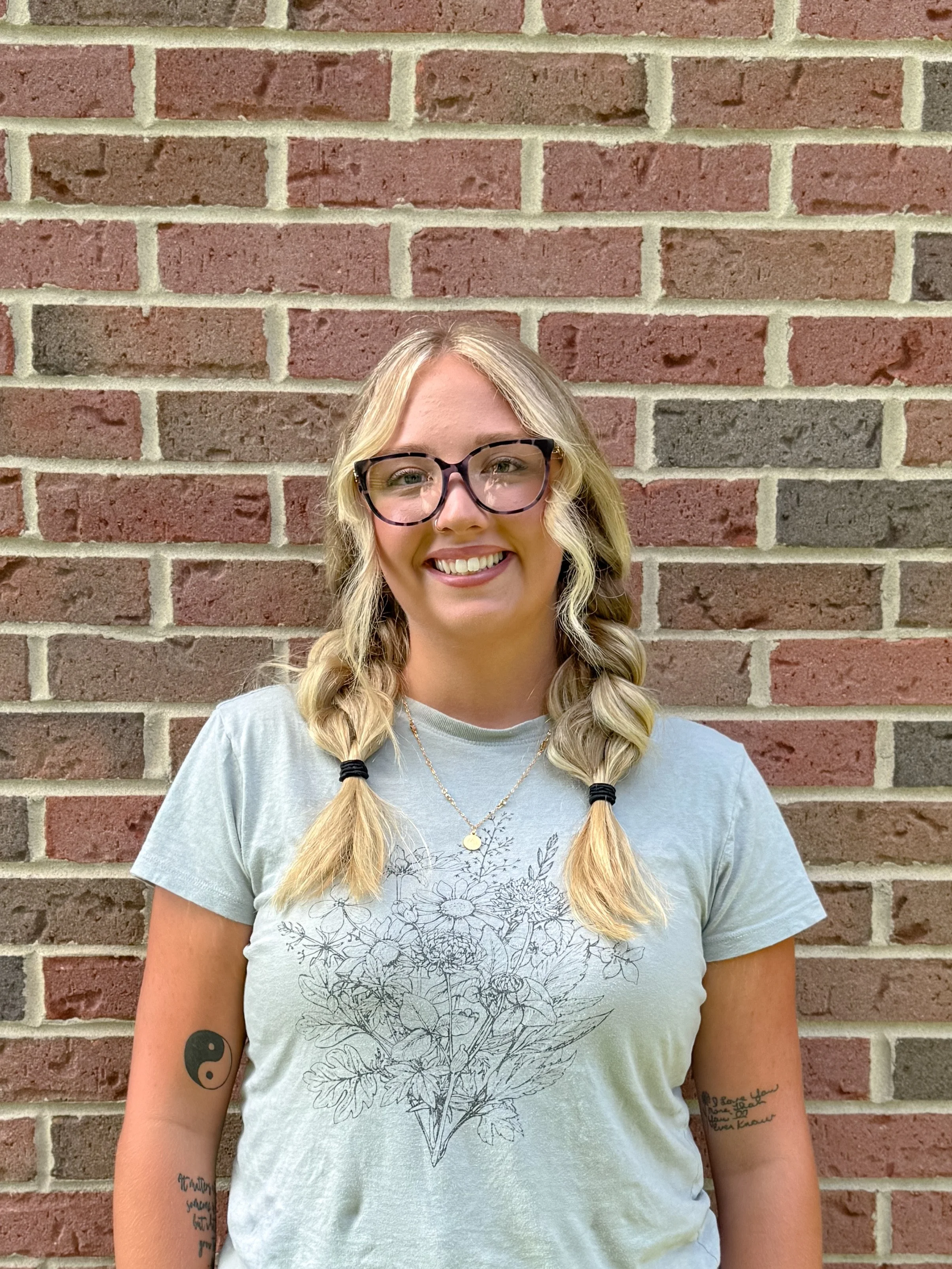 A young woman with blonde hair styled in two braids, wearing glasses, and smiling, stands against a brick wall. She has tattoos on her arms and is wearing a light-colored t-shirt with a floral outline design.