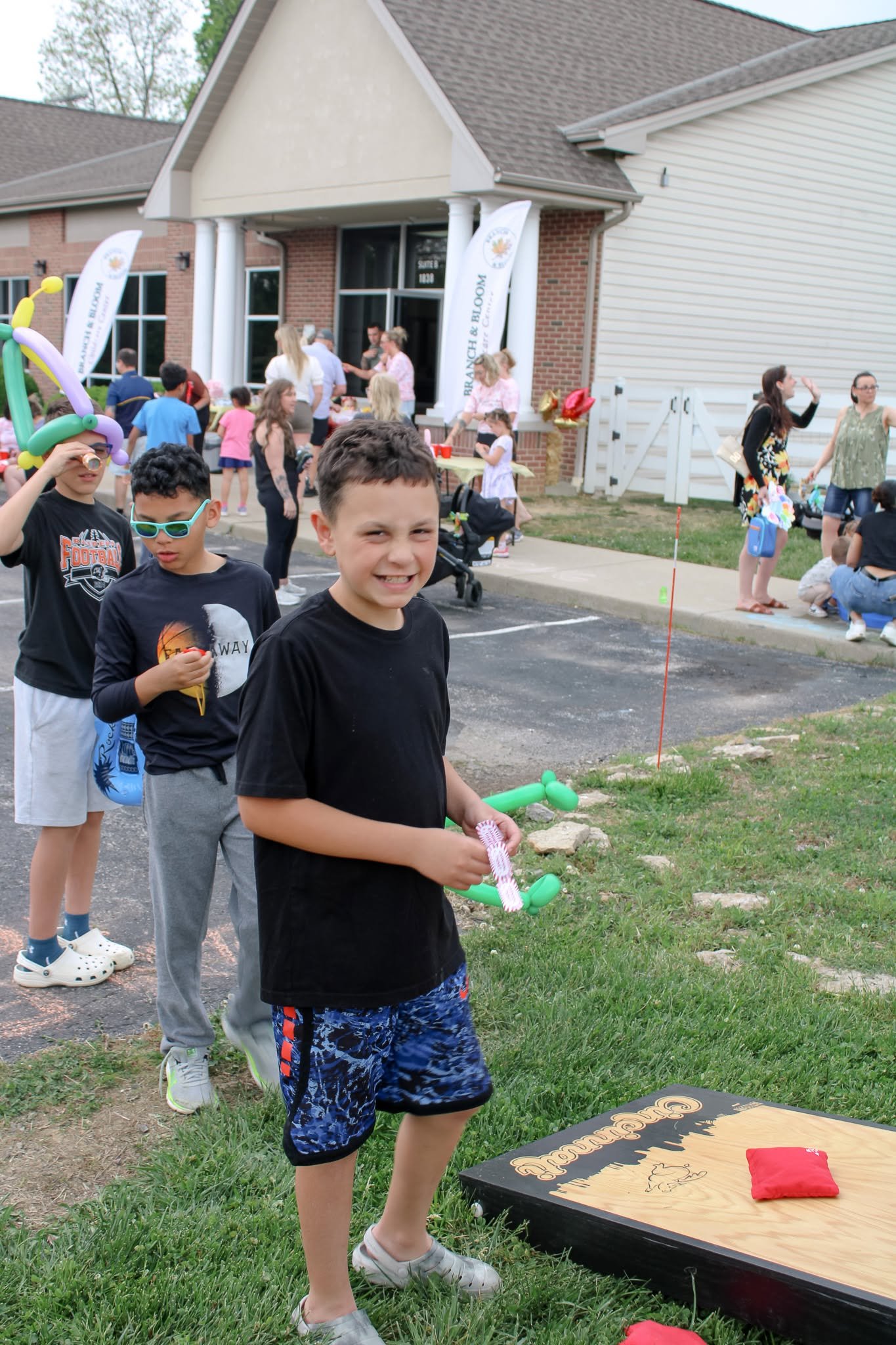 Children and adults playing cornhole outdoors at a social gathering in a residential neighborhood, with a house in the background, balloons, and people socializing.