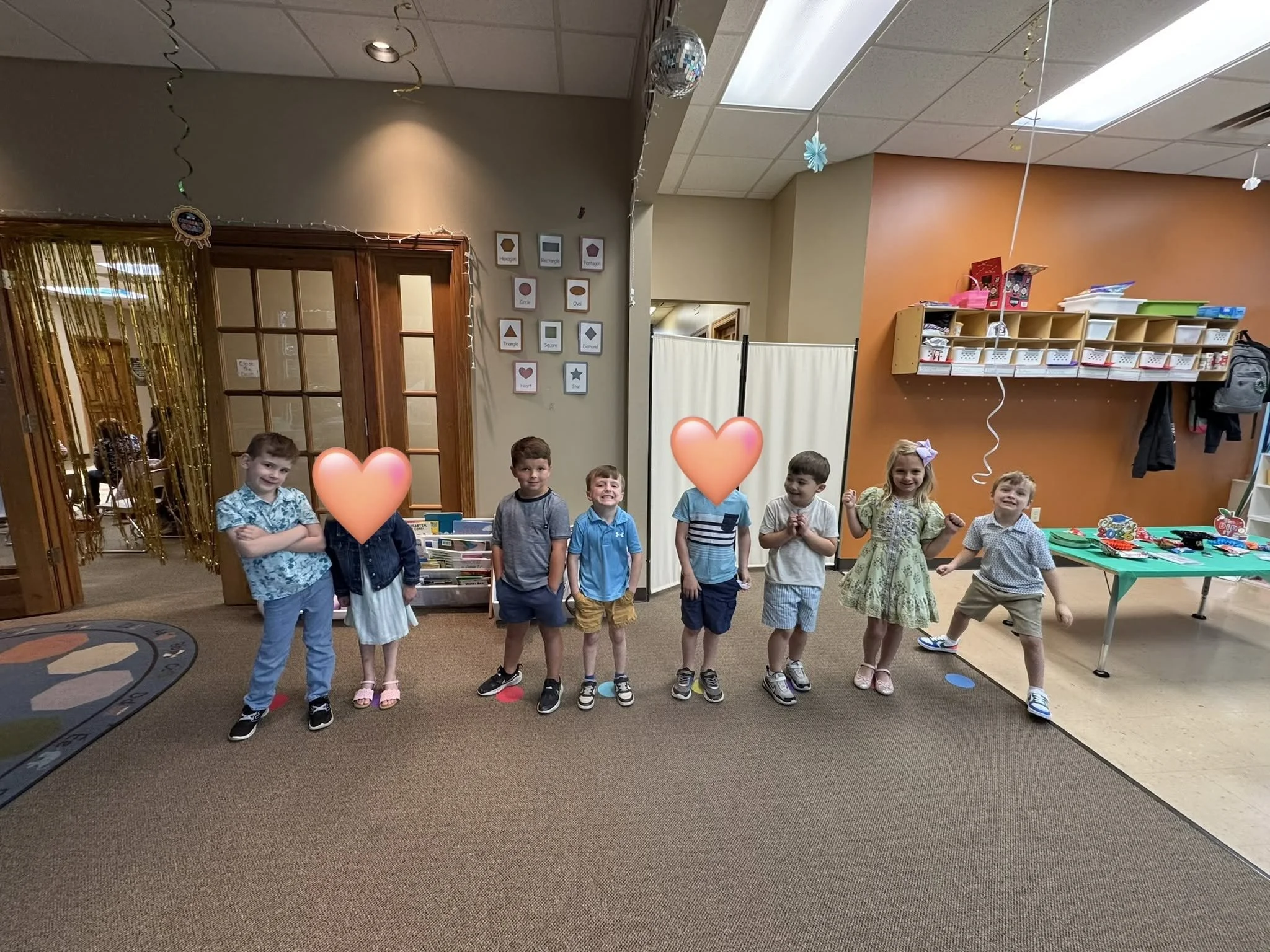 A group of children standing in a classroom decorated for a celebration, with balloons and hearts covering some of their faces, and a table with crafts on it.