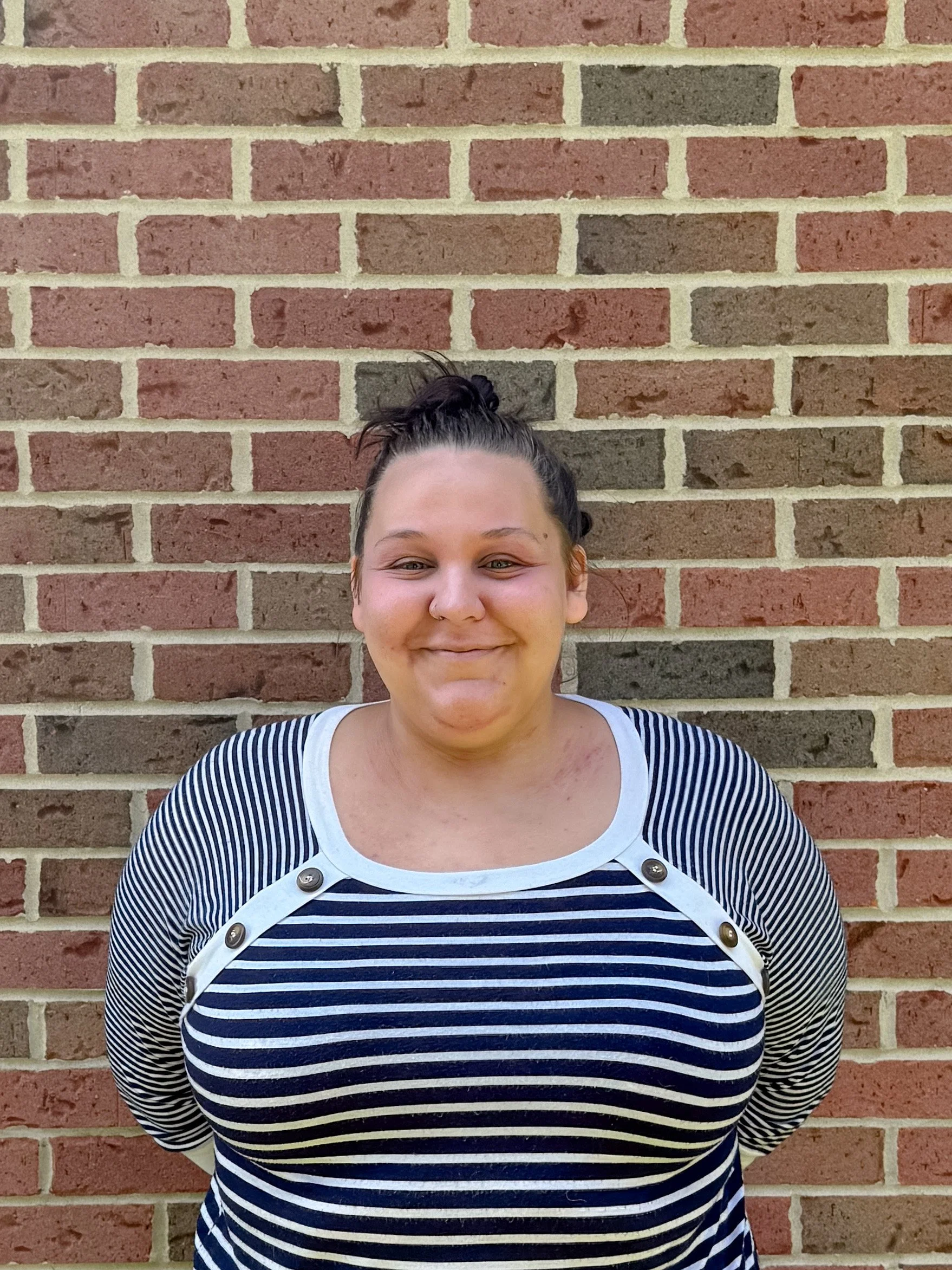A person with dark hair in a bun standing against a brick wall, wearing a striped shirt with buttons on the shoulders.