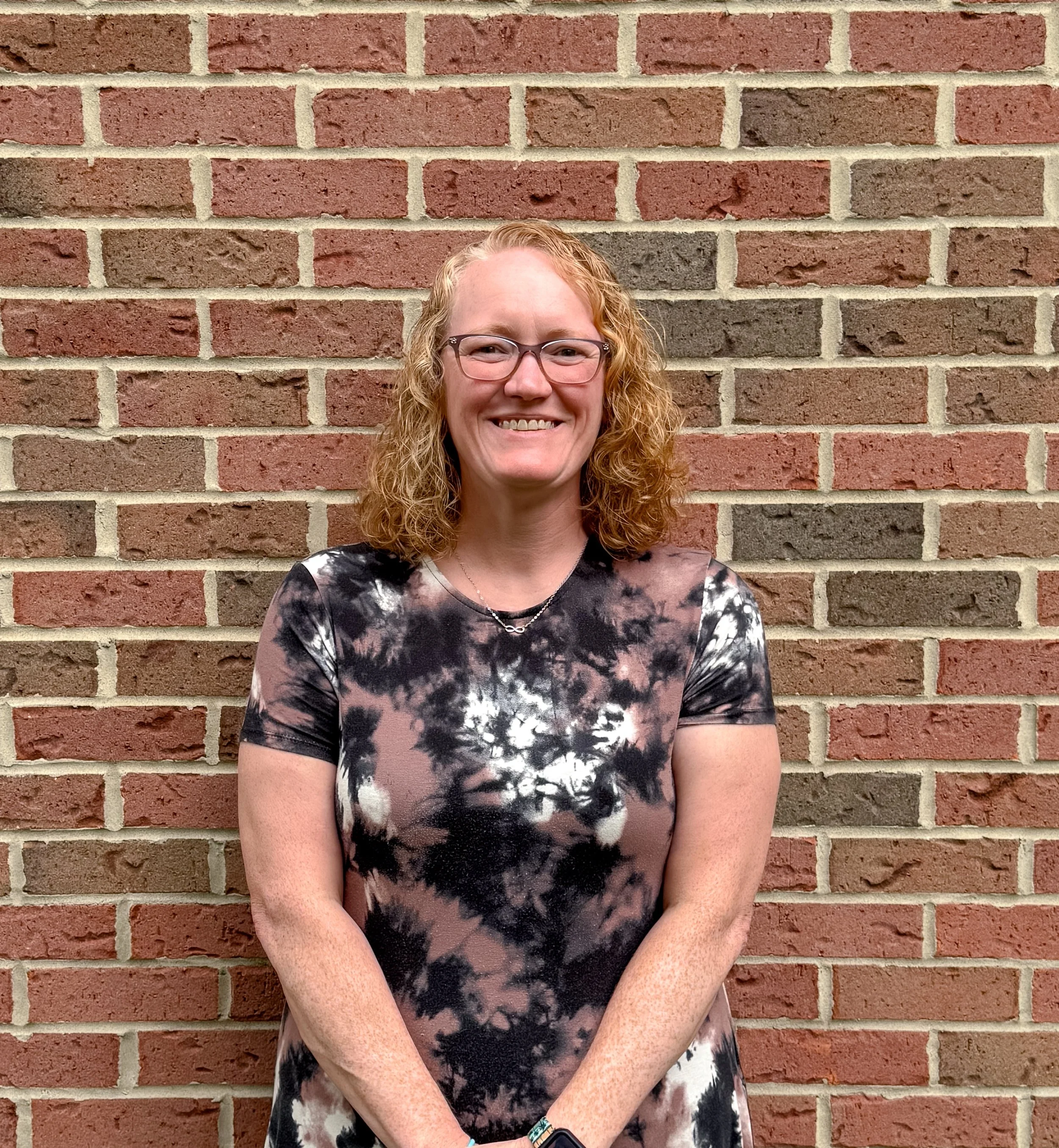 A woman with curly red hair, glasses, and a tie-dye shirt standing in front of a brick wall. She is smiling and has her hands folded in front of her.