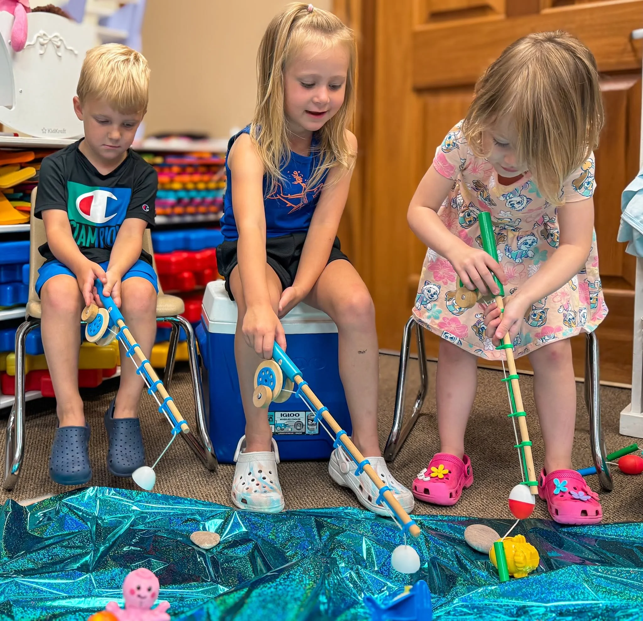 Three children, two girls and one boy, are playing on a pretend outdoor scene with toys and a reflective blue surface, possibly representing water.