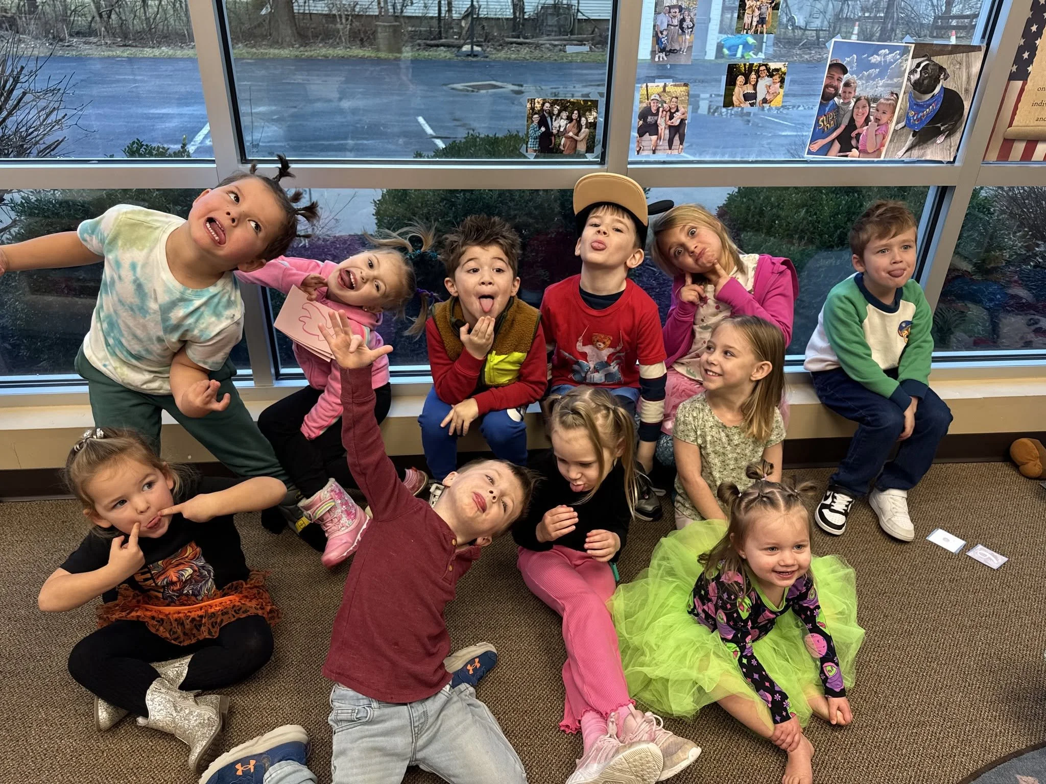 A group of young children posing together indoors, some making silly faces or gestures. Behind them, many photos are taped to a large window, including images of people, dogs, and outdoor scenes.