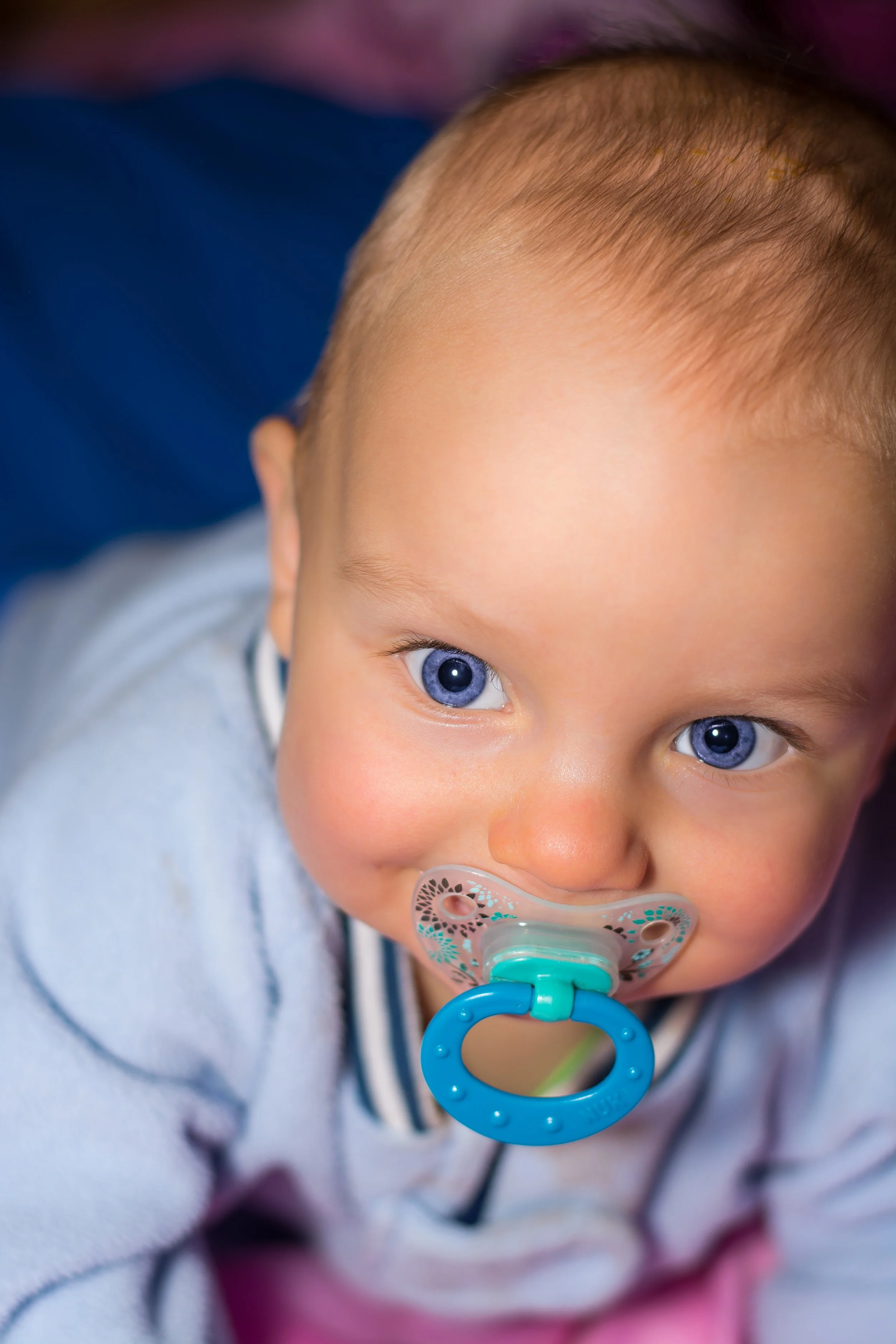 Close-up of a young child's face, showing bright blue eyes, a pacifier in their mouth, and short light brown hair. The child is wearing a gray top with dark and light stripes visible.