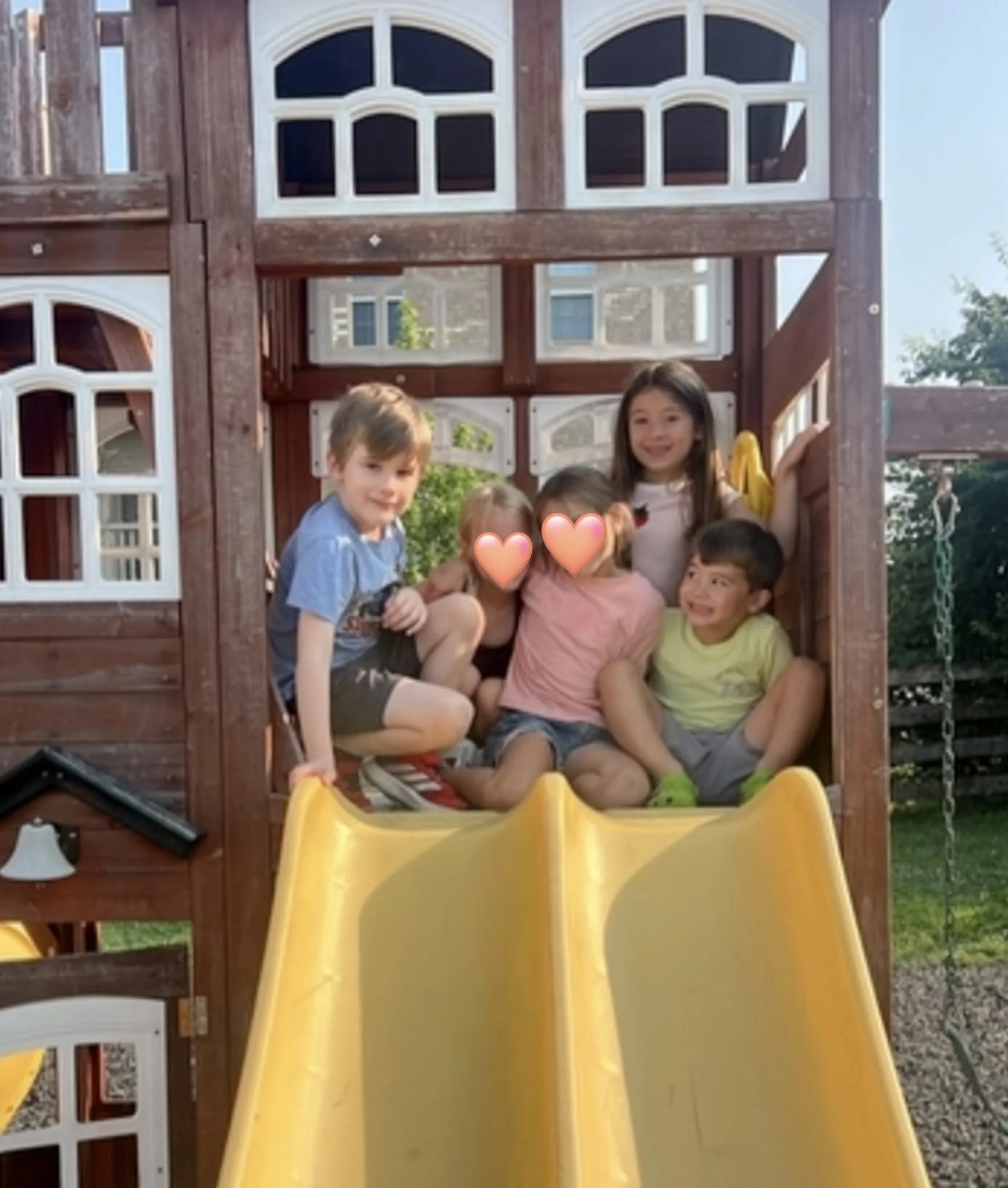 Children playing on a yellow slide attached to a wooden playhouse structure outdoors, with windows and trees visible in the background.