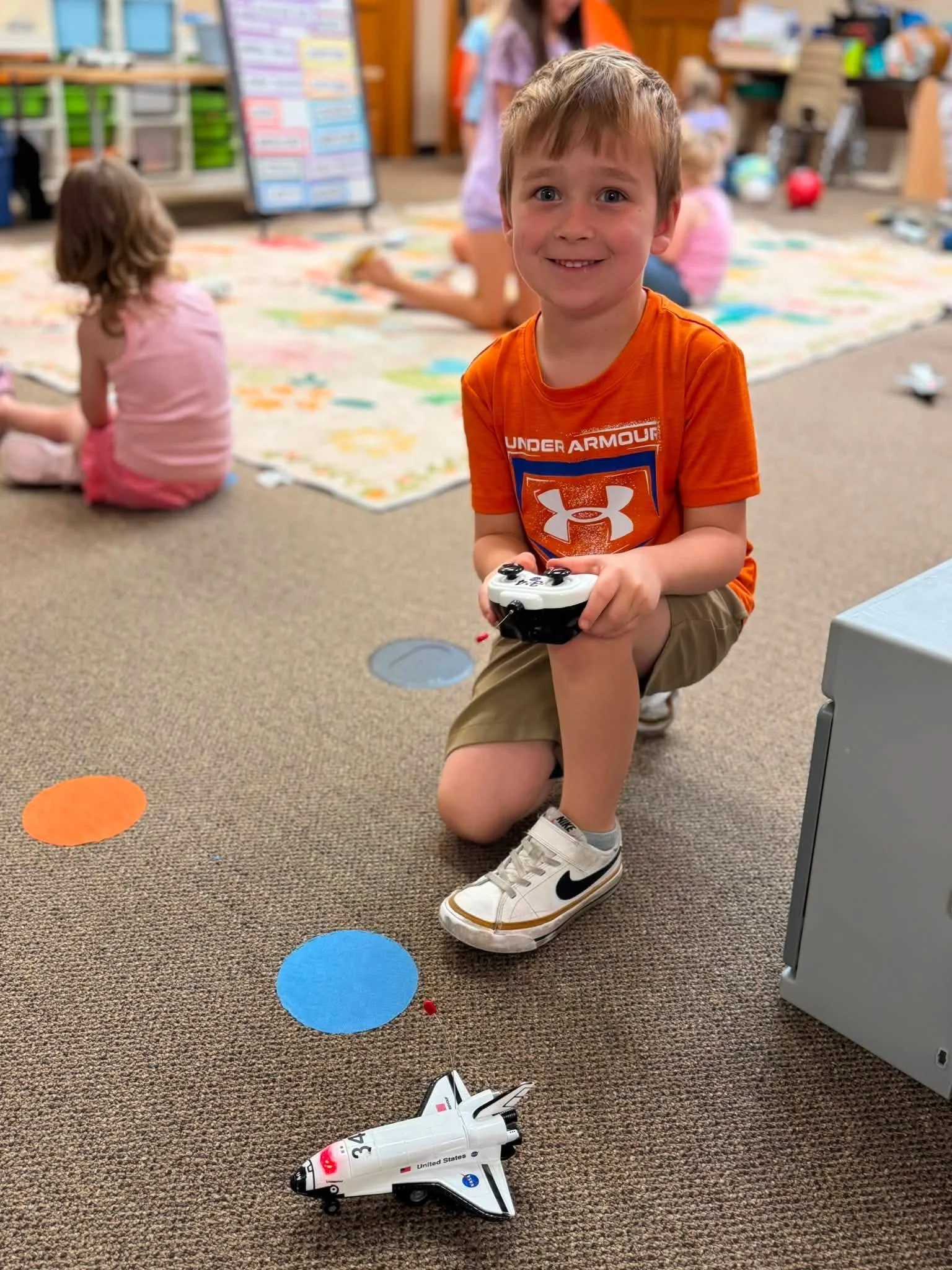 A young boy wearing a bright orange Under Armour shirt, khaki shorts, and Nike sneakers kneels on a carpeted floor in a classroom, holding a toy space shuttle. In the background, several children are sitting or kneeling on a colorful rug with alphabet and number symbols. There are classroom shelves, a whiteboard, and other educational items visible.