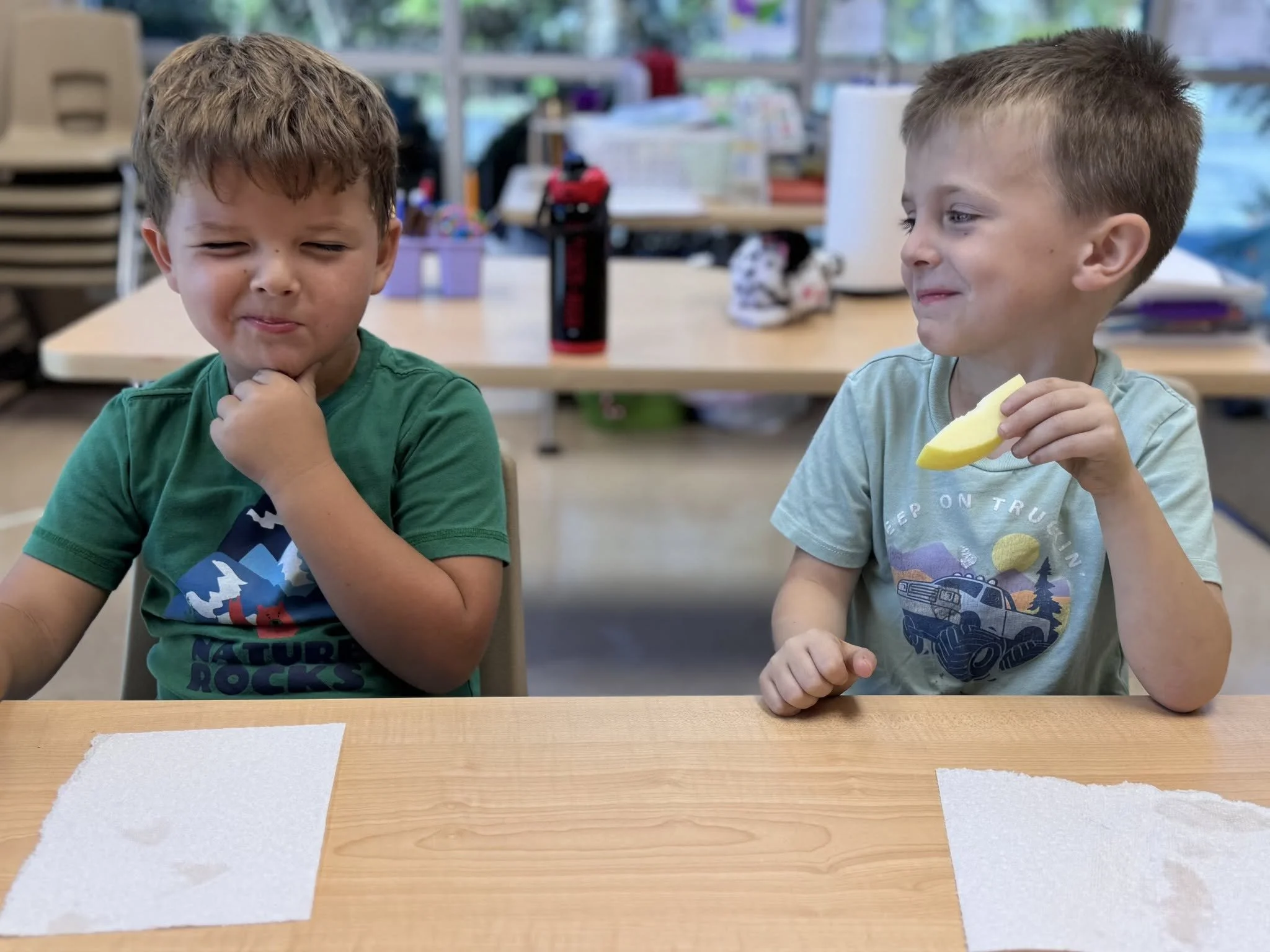 Two young boys sitting at a table in a classroom, one with brown wavy hair and a green shirt, the other with short blond hair and a light gray shirt, holding a slice of yellow fruit.