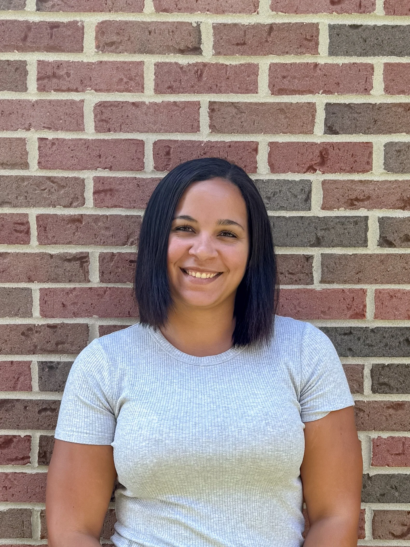 A woman with black hair smiling, wearing a light gray shirt, standing against a brick wall.
