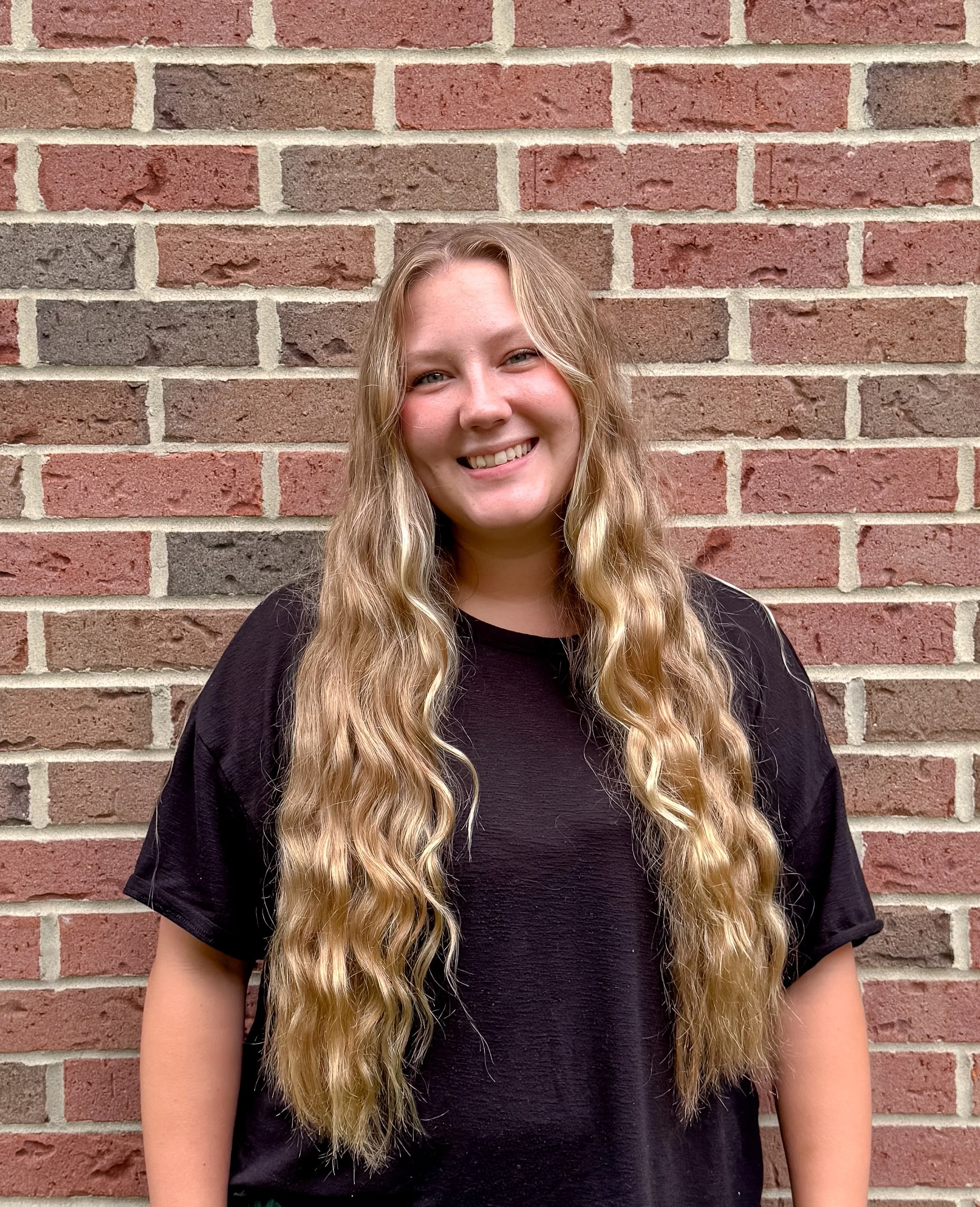 A young woman with long, wavy blonde hair smiling, standing against a brick wall background.