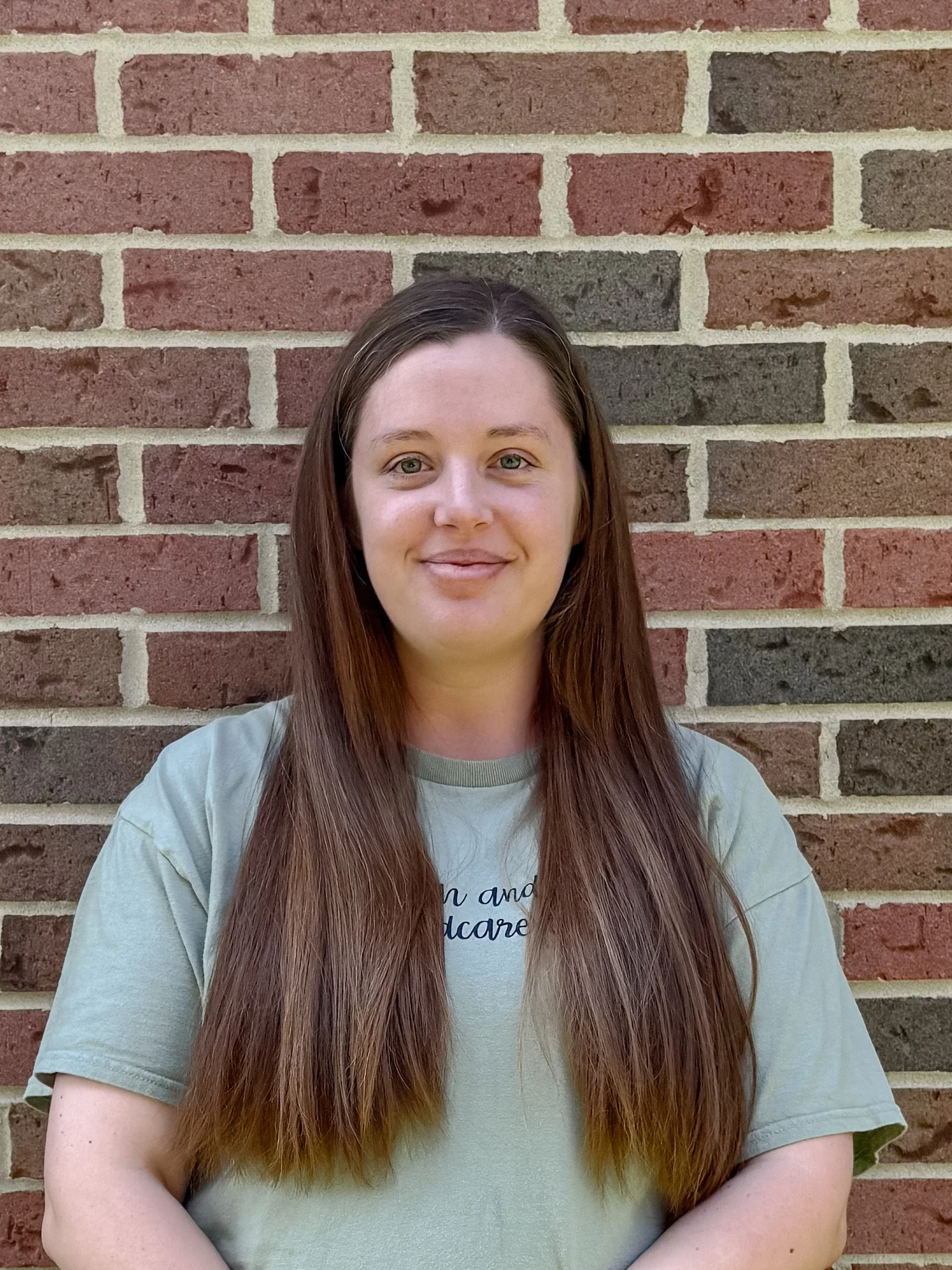 A woman with long brown hair, wearing a light gray T-shirt, standing in front of a red brick wall with a smile.