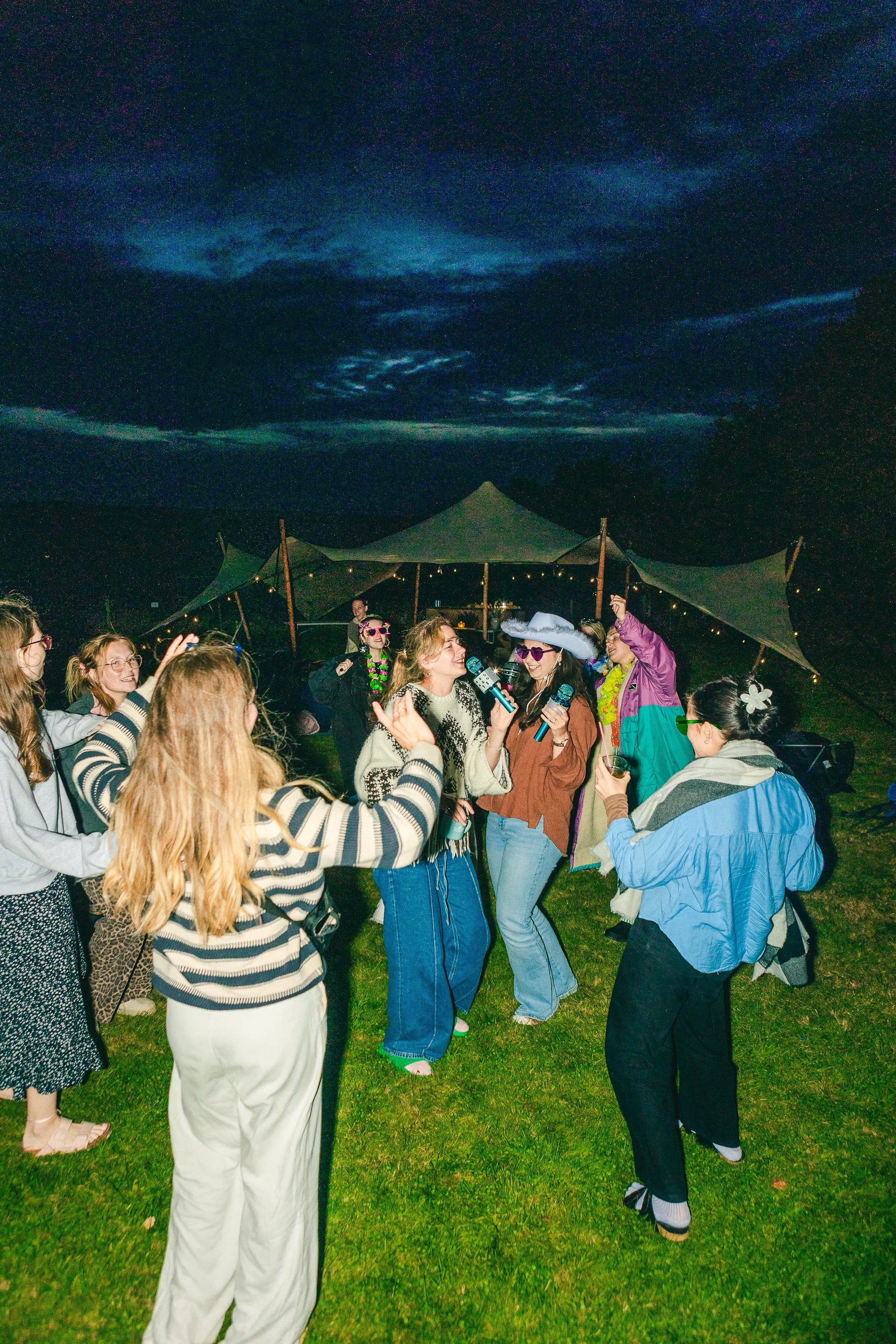 A group of people dancing and singing together outdoors at night near a large tent, some wearing colorful or retro clothing and accessories, with a dark sky overhead.