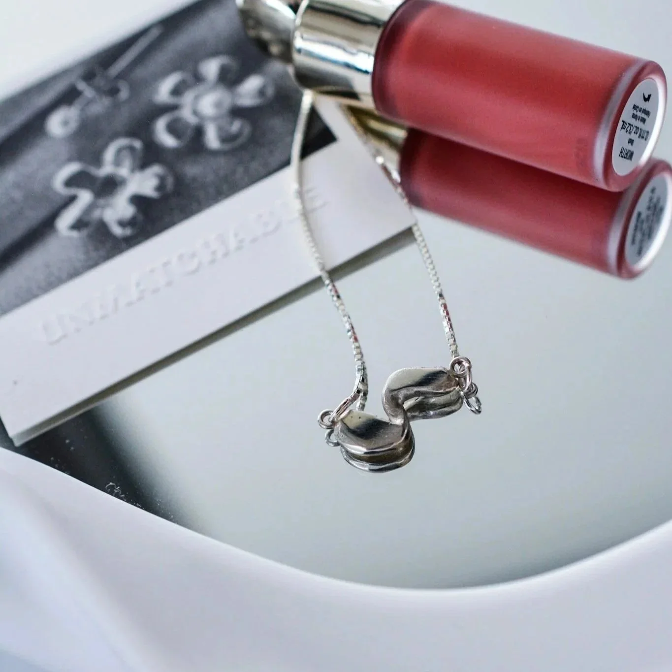A silver necklace with a swan-shaped pendant resting on a mirror surface, with a black and white jewelry box and two red lipstick tubes nearby.
