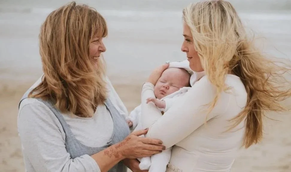 Two women holding a sleeping baby on a beach, smiling at each other.