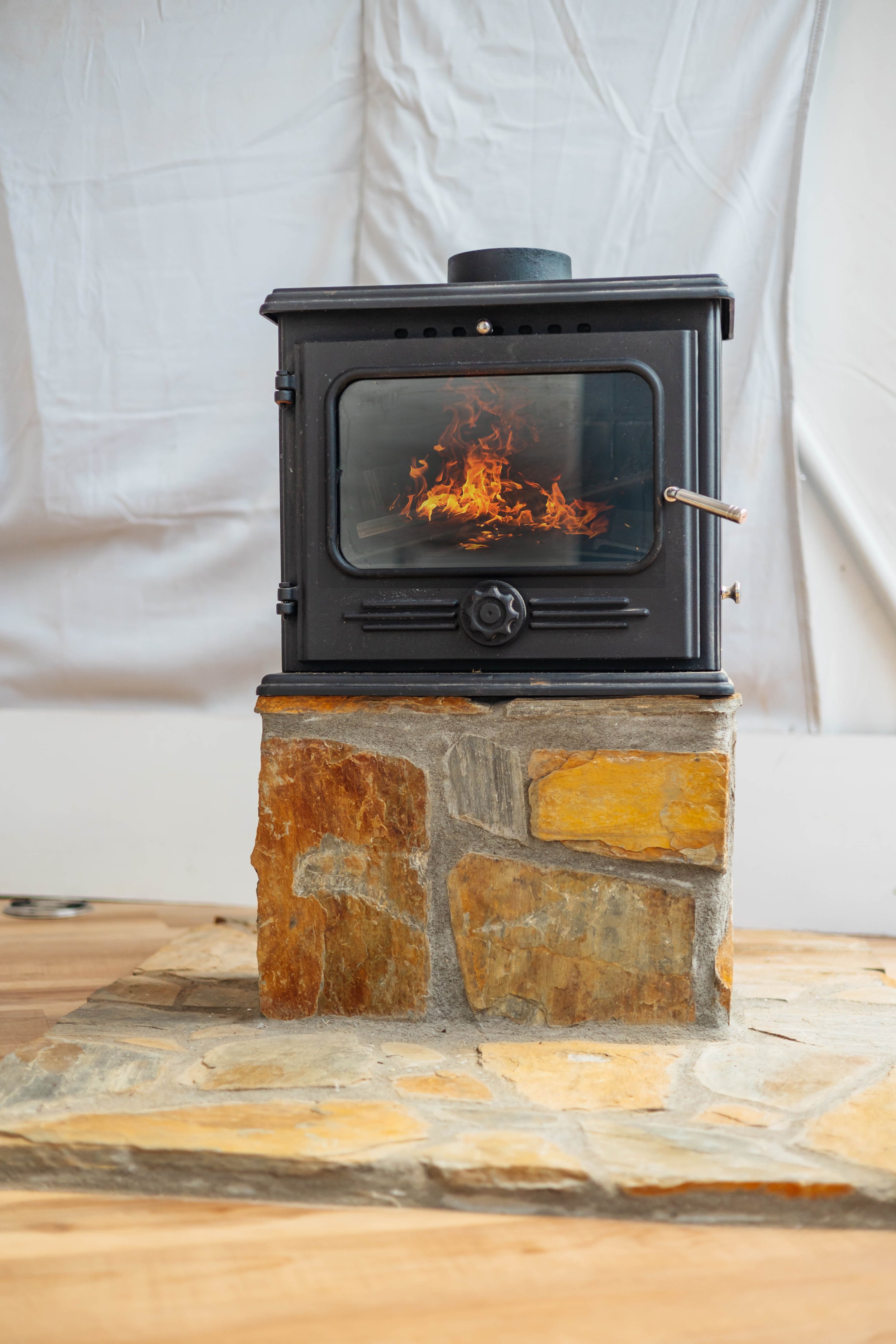 A black cast iron wood stove with a glass door showing flames inside, sitting on a stone base, on a wooden floor, with a white fabric backdrop.