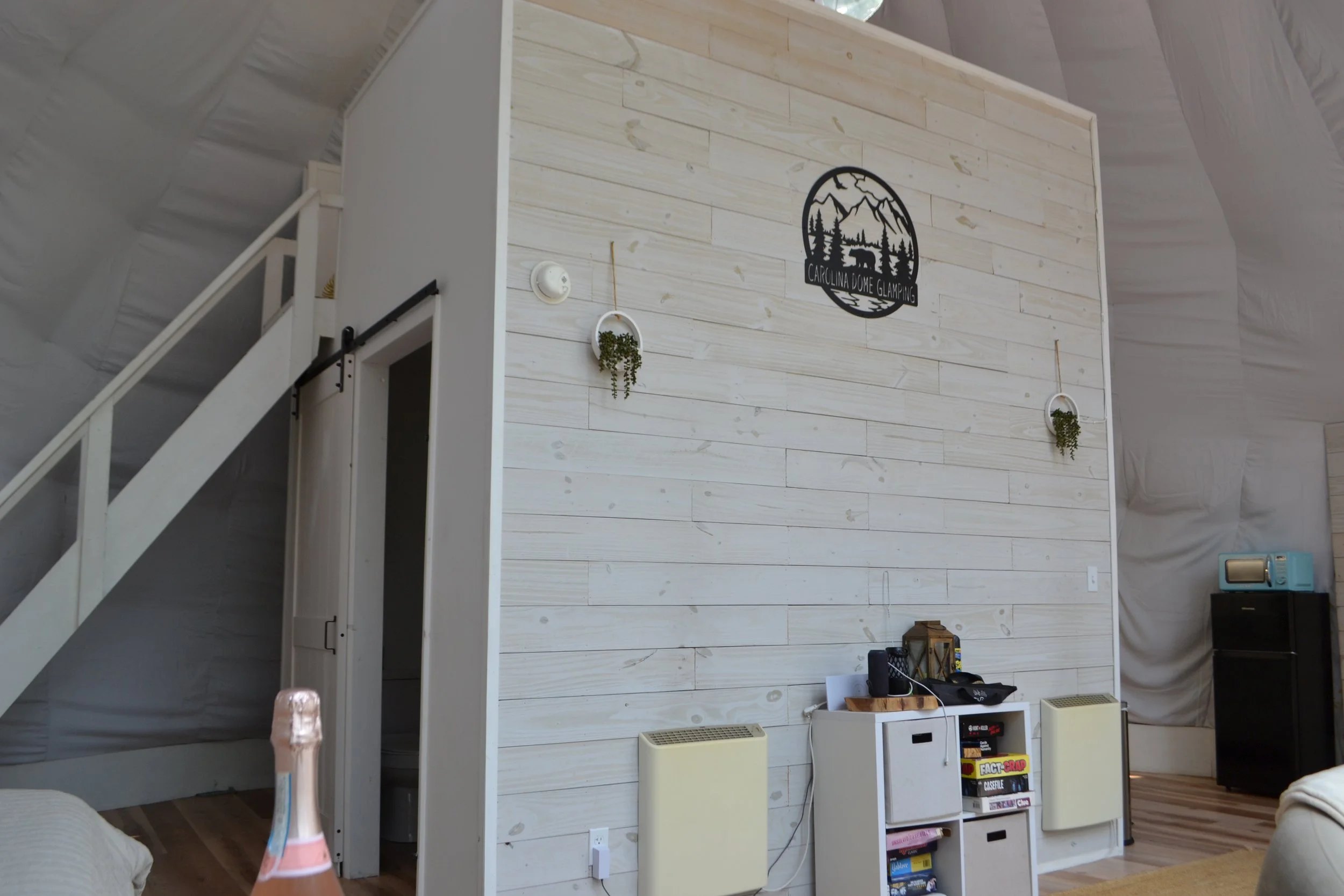 Interior of a room with a white wooden wall and a black circular wall art that says 'Carolina DOME Glamping' with mountains and trees. There are two small hanging planters with greenery on the wall. Below, there are two small yellow and white speaker