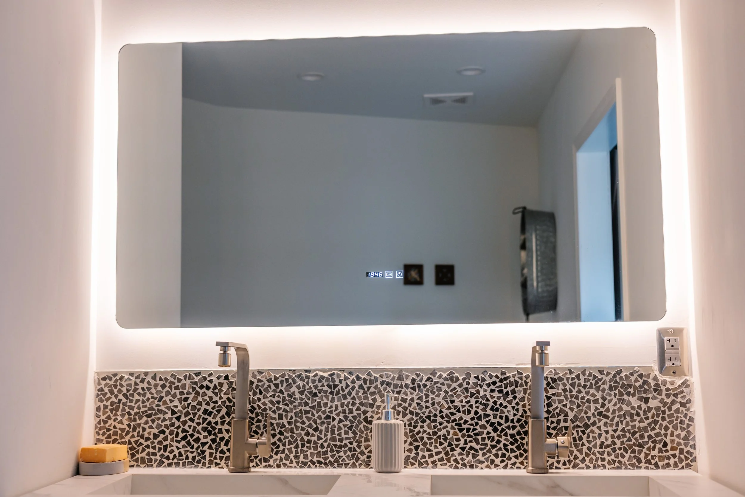Modern bathroom double sink vanity with mosaic tile backsplash, square soap dispenser, soap, and large backlit mirror.