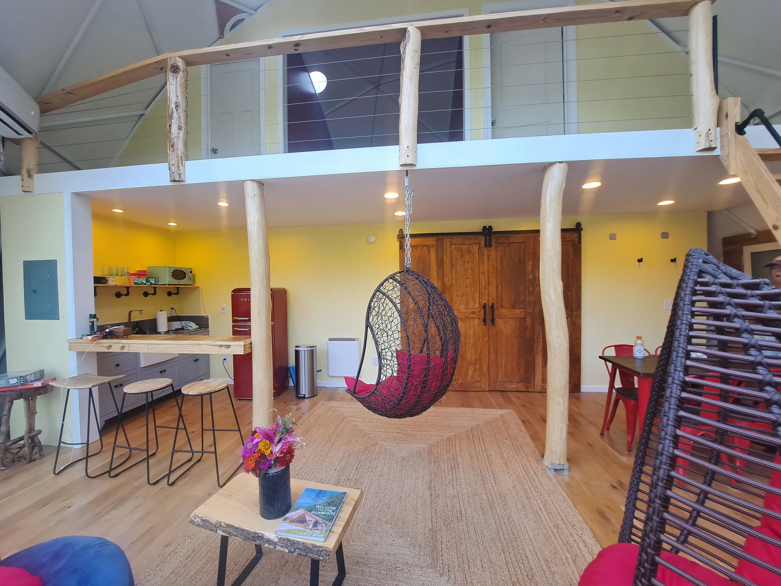 Interior view of a cozy space with a loft, hanging chair, small kitchen area, table, and chairs, with natural wood accents and modern decor.