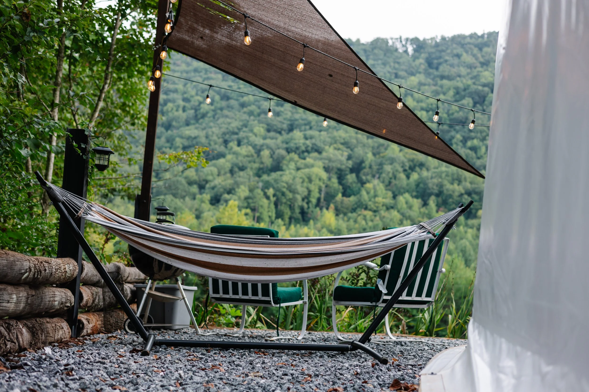 A cozy outdoor seating area with a hammock, two chairs with green cushions, string lights overhead, surrounded by lush green trees and overlooking a hillside.