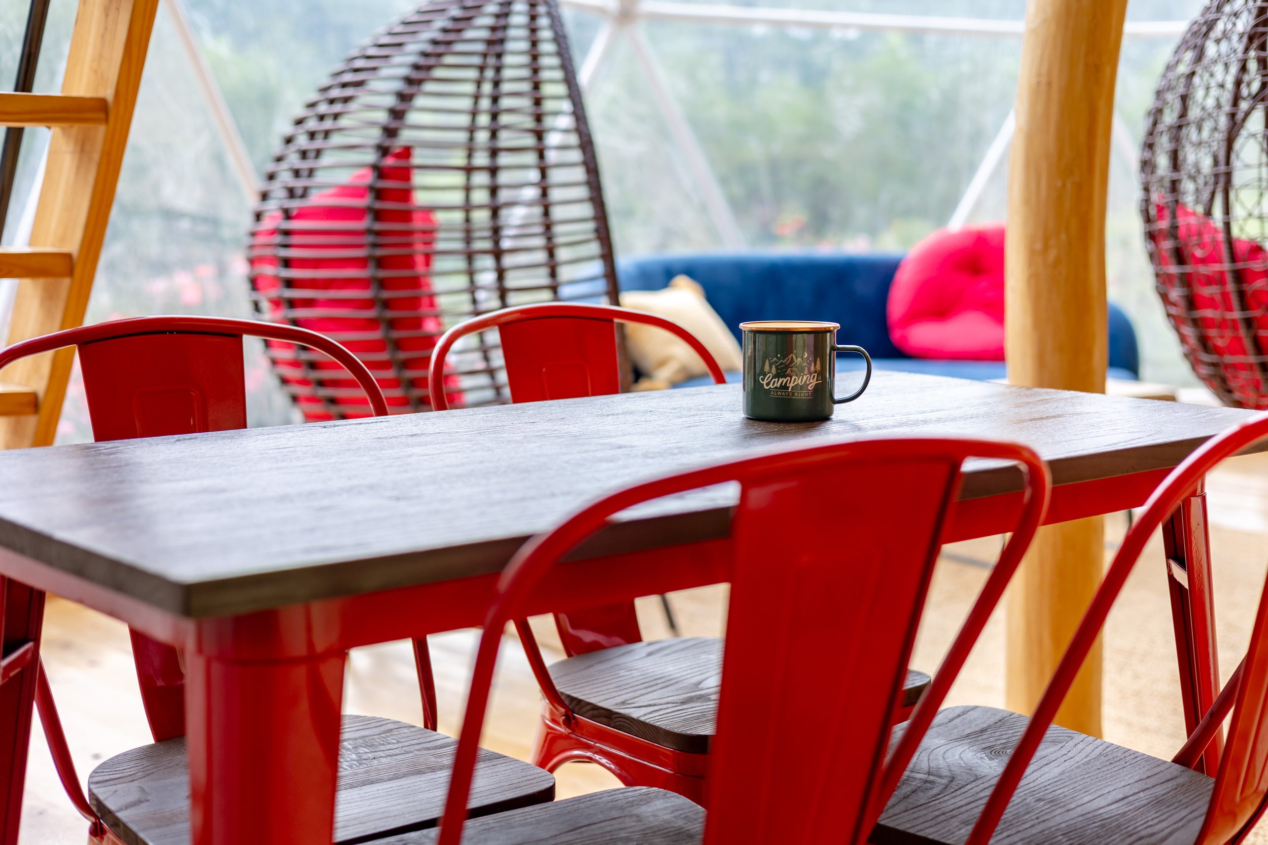 A wooden table with red metal chairs, a coffee mug, and hanging egg-shaped wicker chairs with cushions in the background.