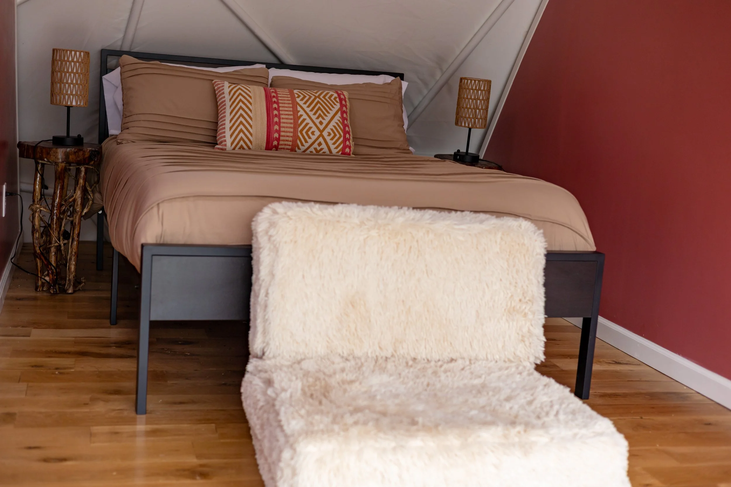 A bedroom with a bed, beige bedding, and decorative pillows. Two nightstands with wicker lamps, a small rustic table on the left, and a faux fur blanket draped over the foot of the bed.