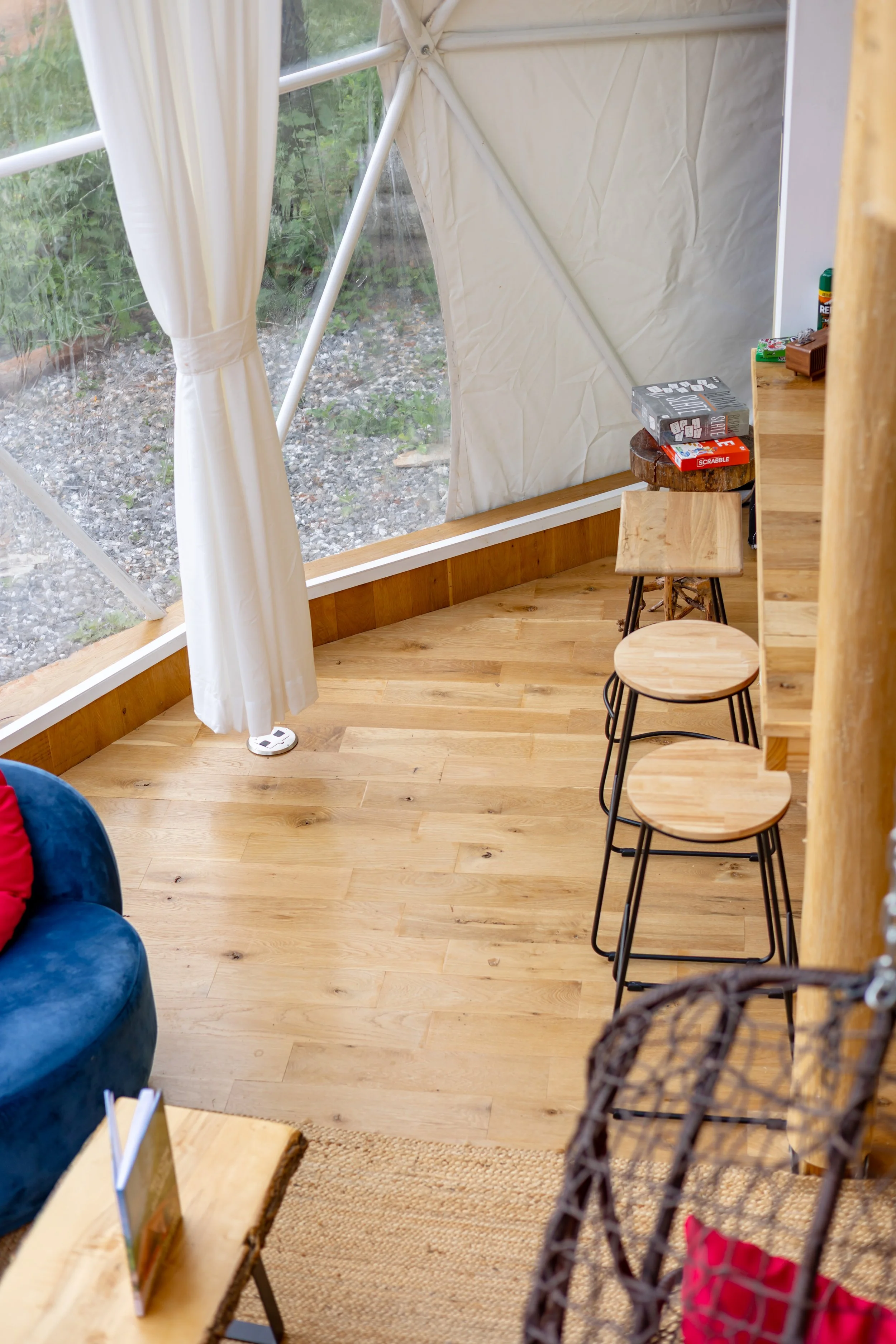 Inside a cozy glamping tent with wooden flooring and furniture, including stools and a wicker chair. Large transparent window with white curtains looking out to a natural outdoor scene. Items like board games and glue on a wooden countertop.