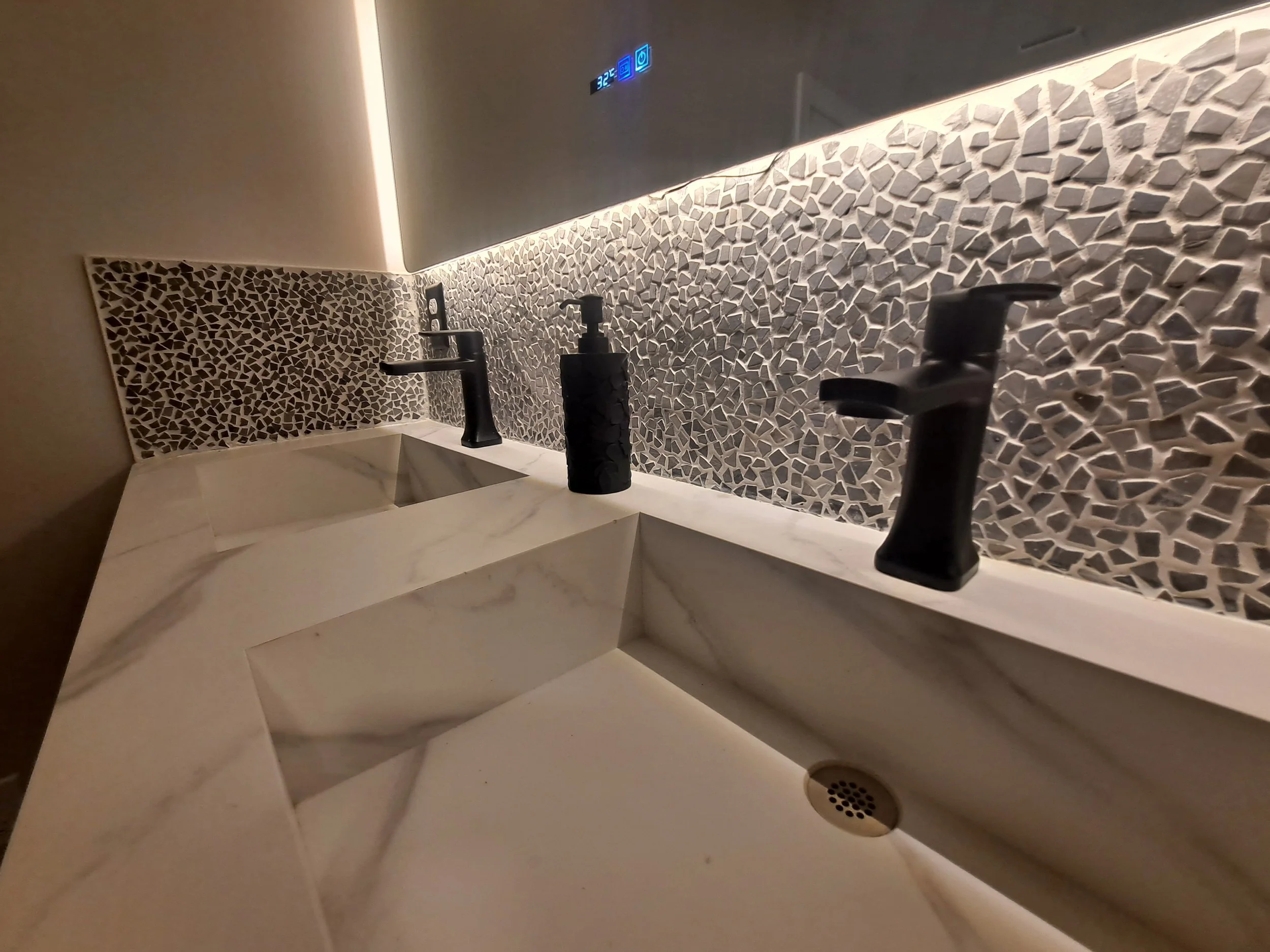 Close-up of a modern double bathroom vanity with white marble sinks, black faucets, and a textured mosaic backsplash, with a digital temperature display above.