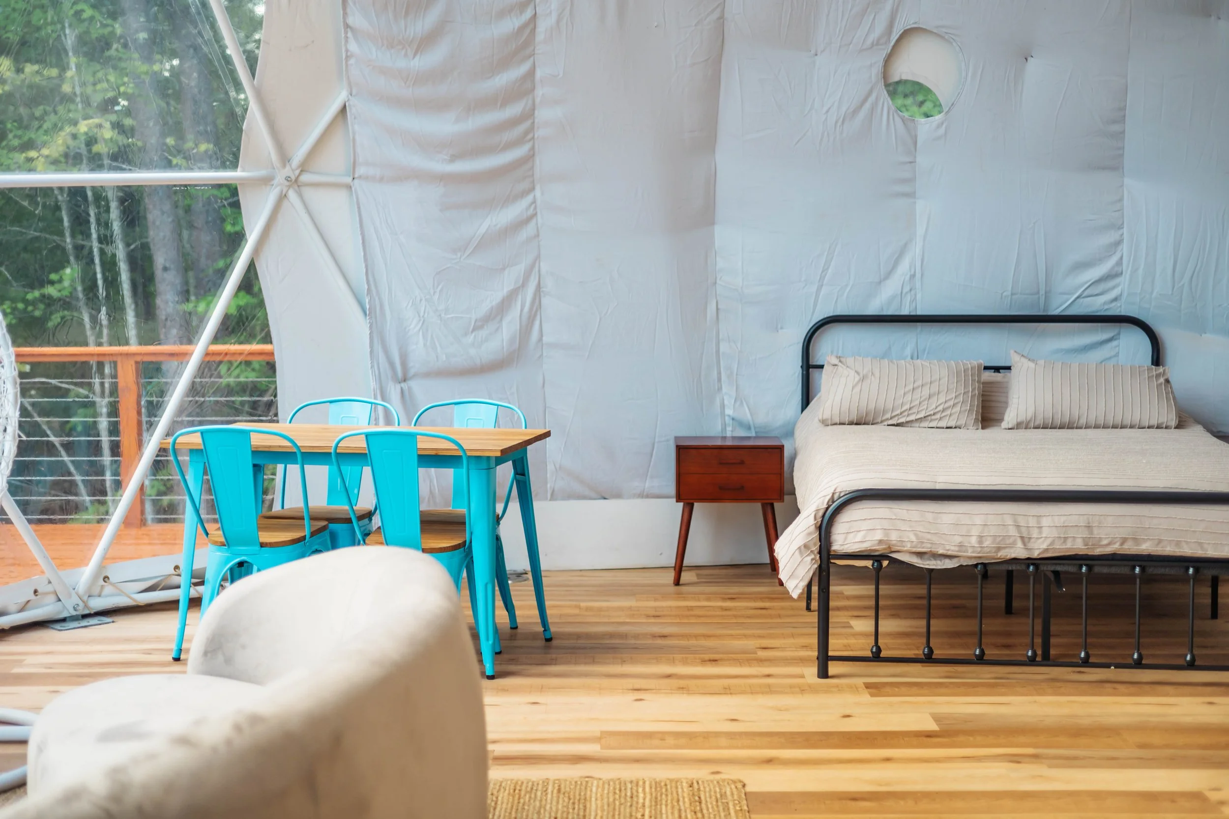 Interior of a cozy bedroom in a glamping tent with a black metal bed frame, beige striped bedsheets, a small wooden nightstand, a wooden dining table with four bright blue metal chairs, and a white plush armchair on a wooden floor, with a circular wi
