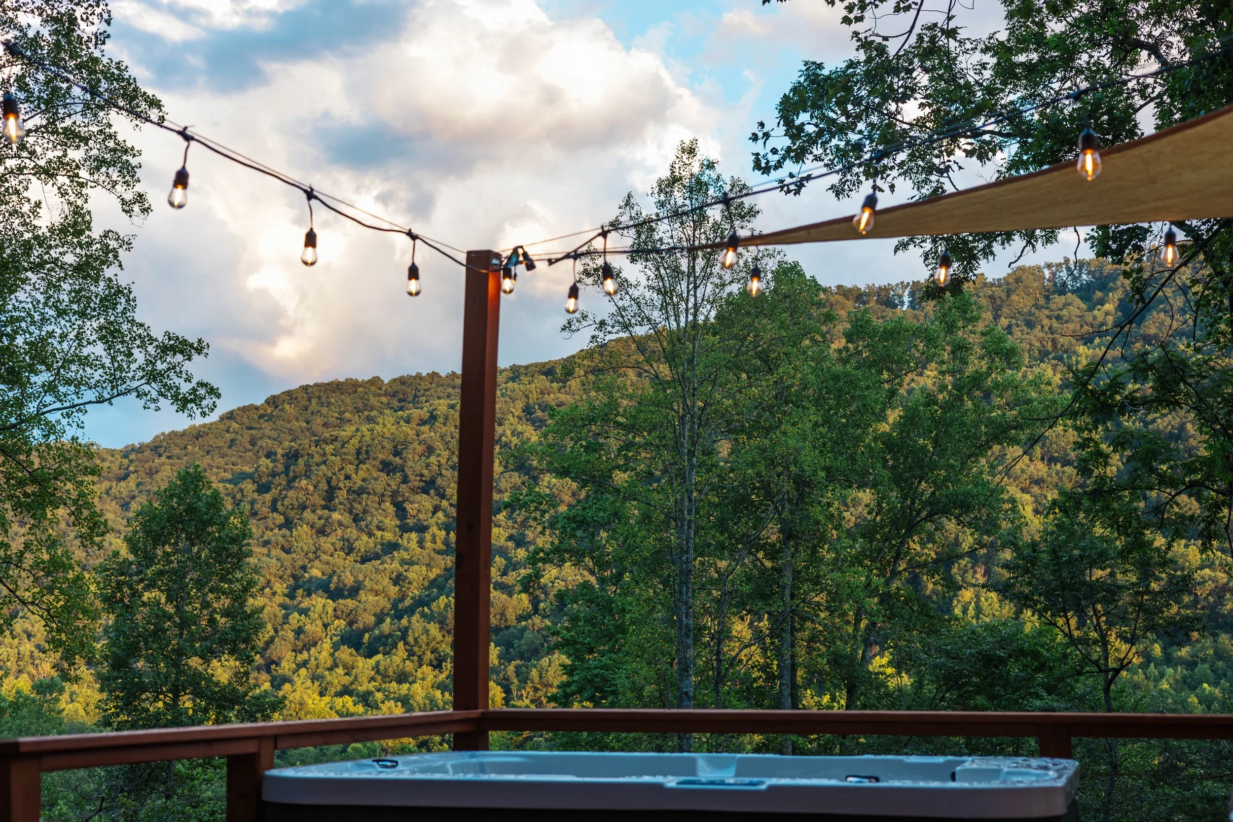 An outdoor deck with string lights and a hot tub, overlooking a green forested hillside during the daytime with partly cloudy skies.