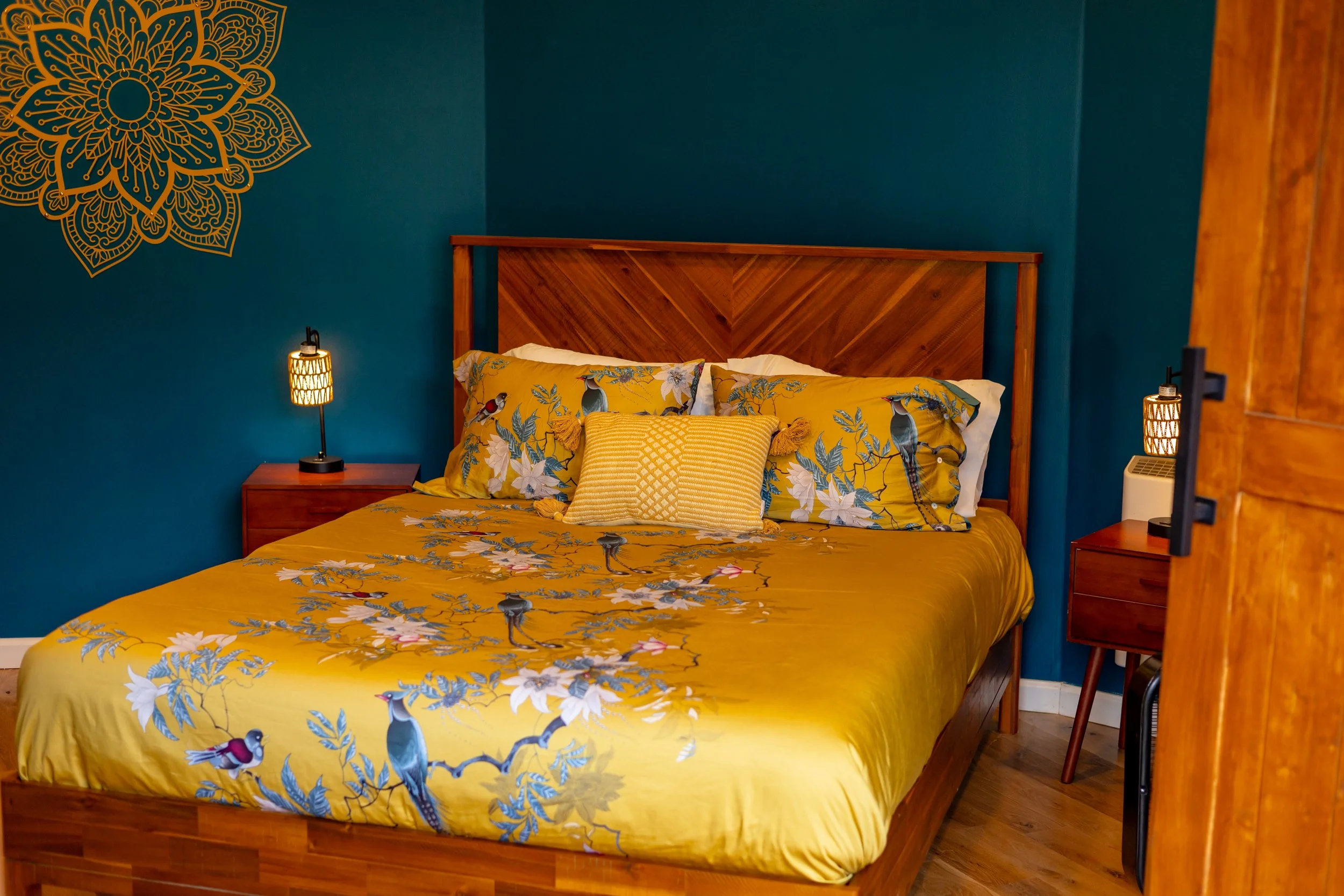 A bedroom with a wooden bed frame, yellow floral and bird patterned bedding, and teal walls with a decorative mandala design in the corner. There are two nightstands with matching lamps on each side of the bed.