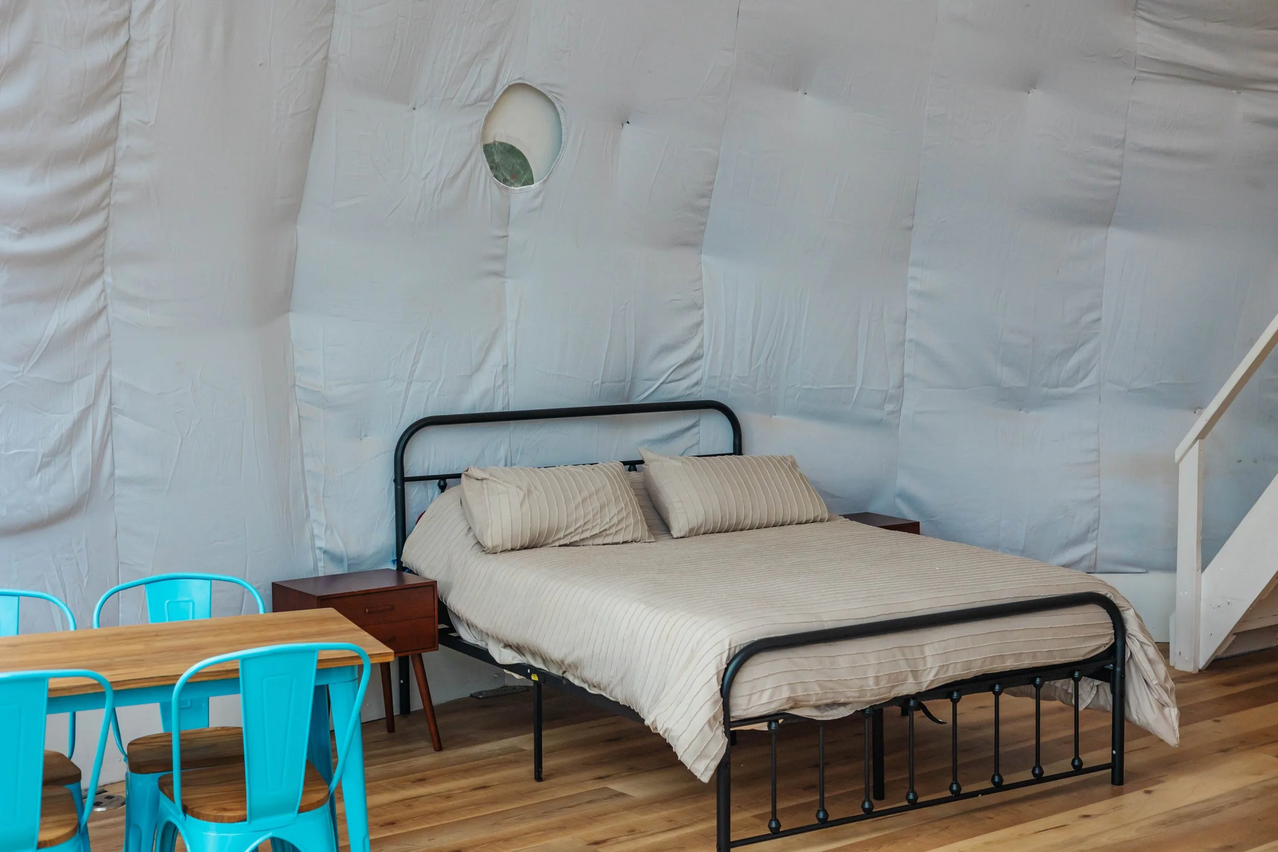 A bedroom with a black metal bed frame, beige striped bedding, two pillows, a wooden nightstand, a wooden dining table, and blue chairs, inside a room with white, padded, insulated walls.